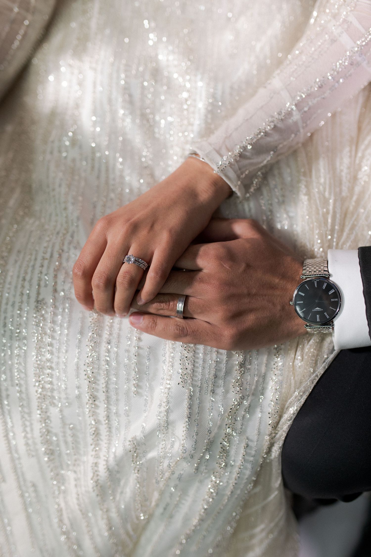 Close up of wedding rings and hands with a watch against a sparkly white dress fabric.