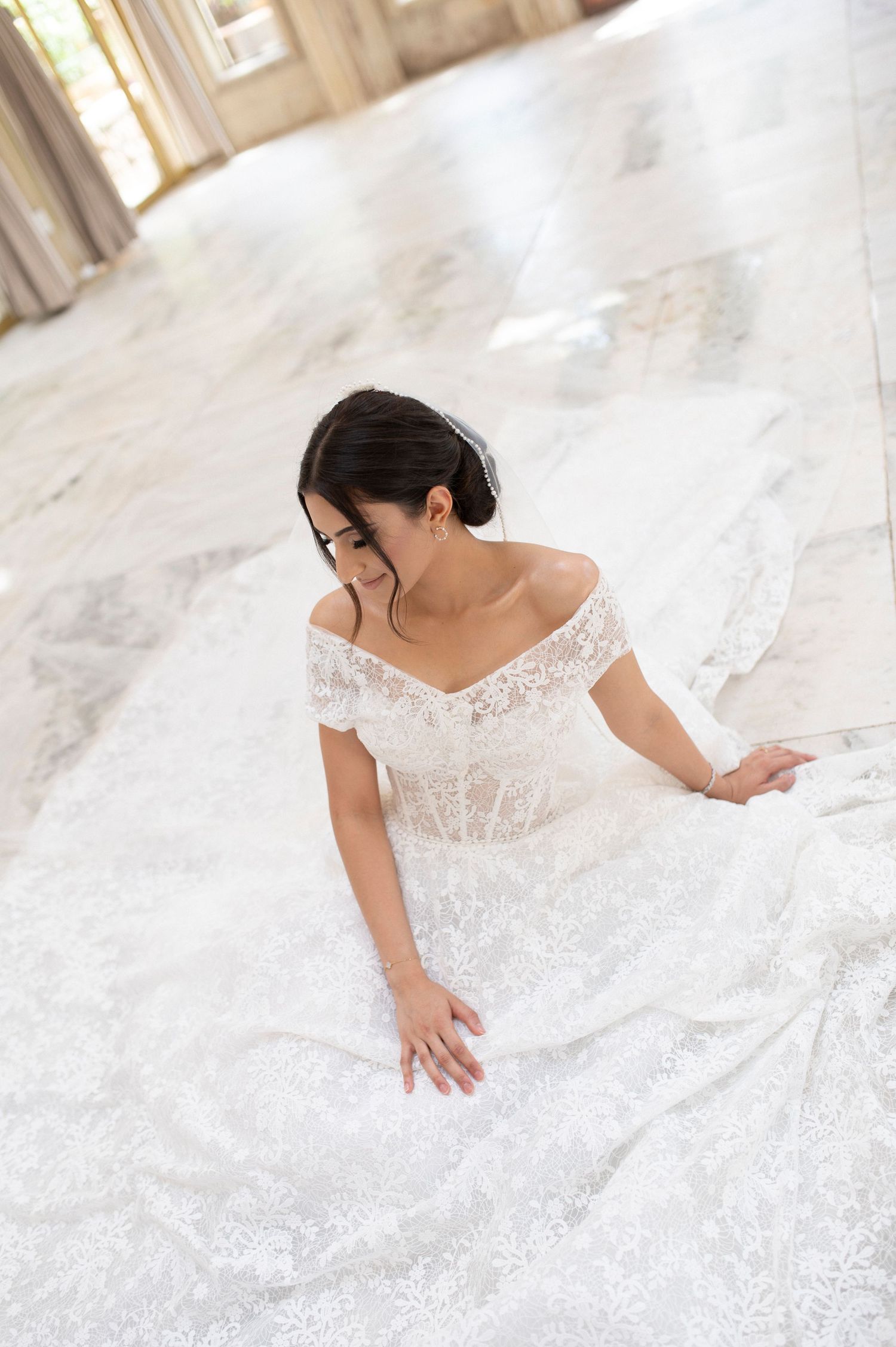 Close up of bride in off-shoulder lace wedding dress with intricate details sitting on marble floor.