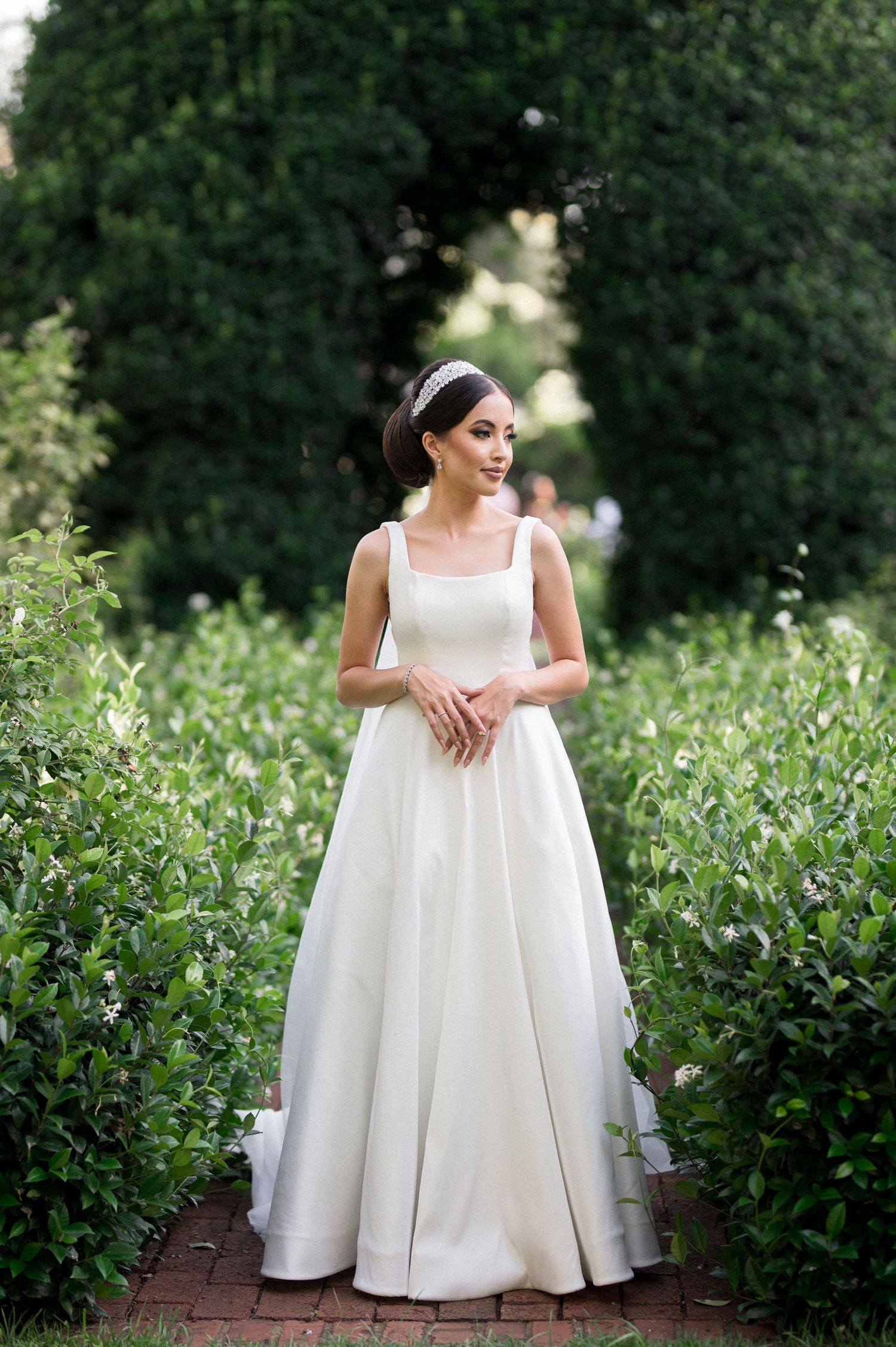 A bride in an elegant simple white wedding dress poses in a garden setting.