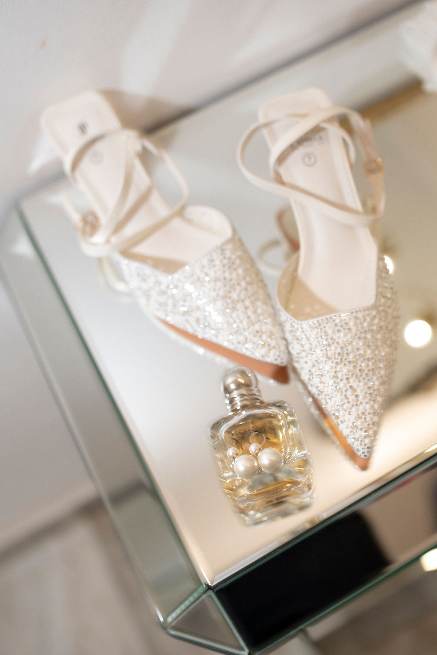 Sparkly bridal shoes and perfume bottle displayed on mirrored surface.