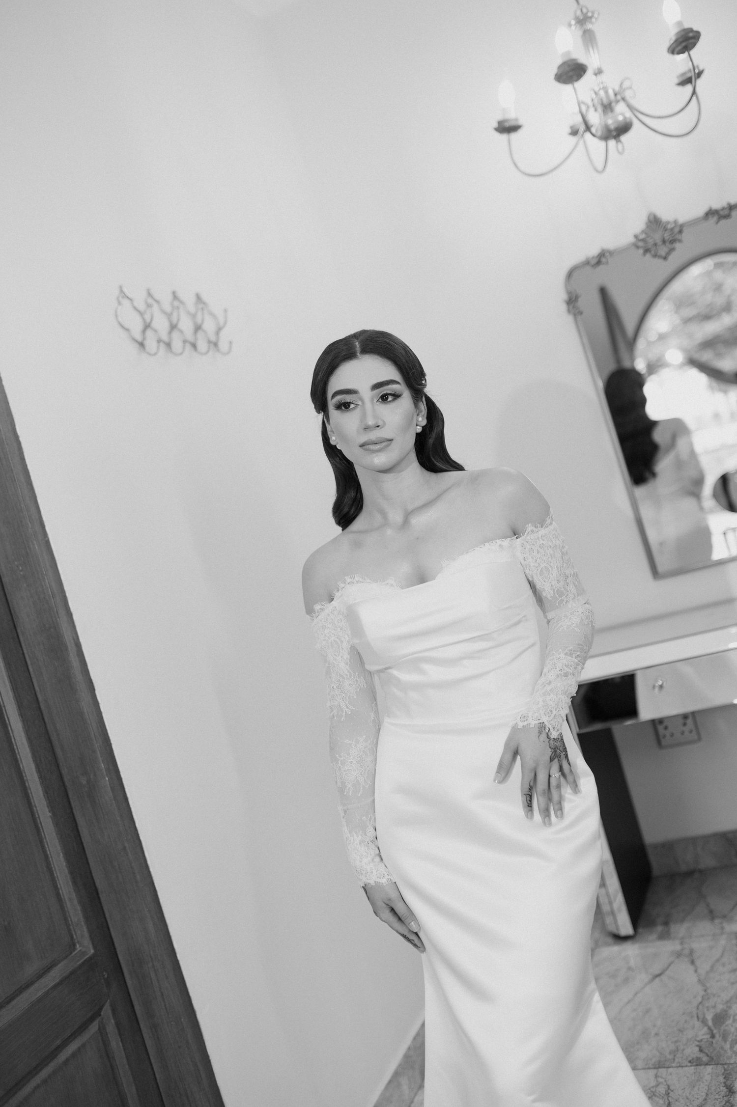 Black and white shot of bride in fitted off-shoulder wedding gown posing in elegant room setting.