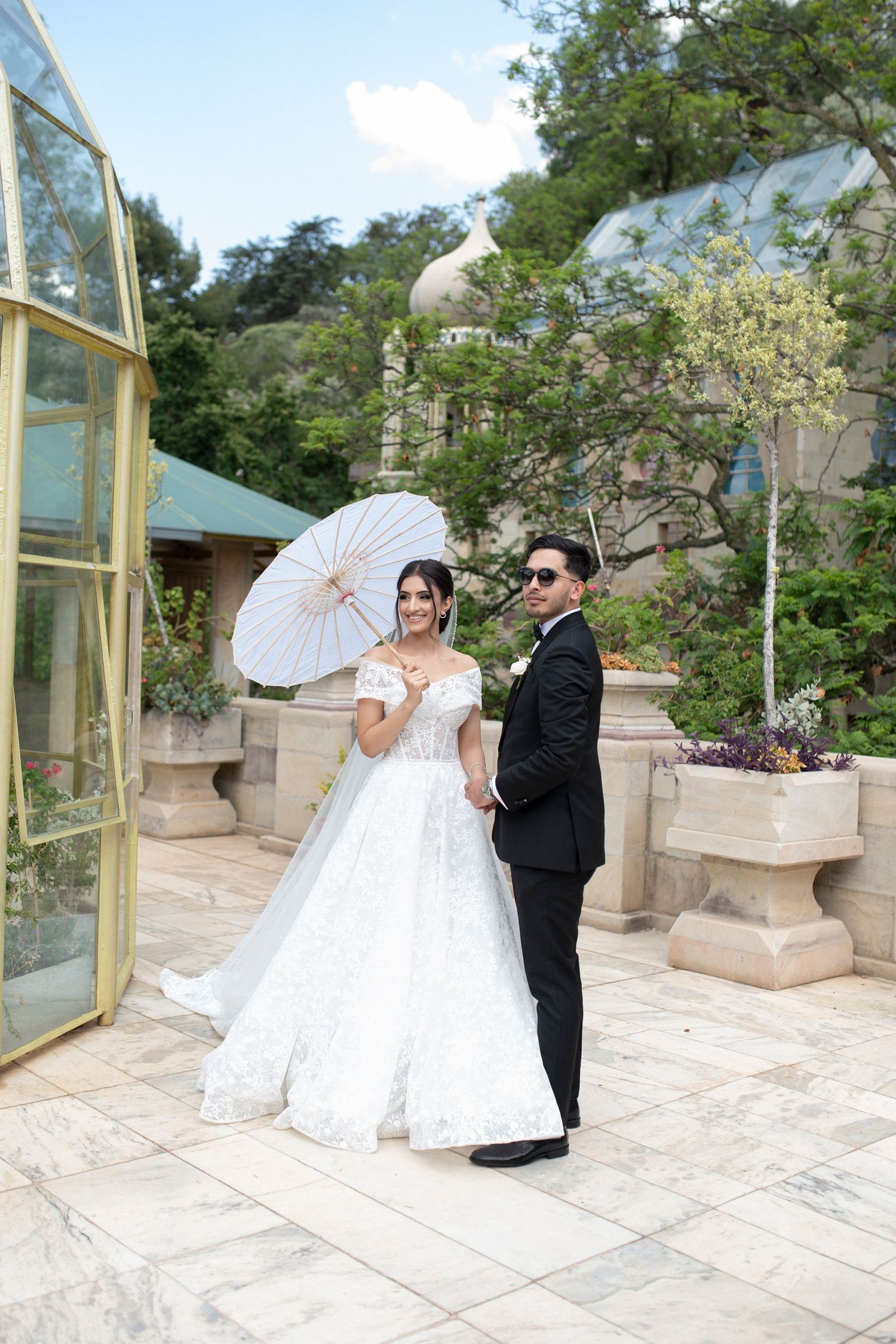 Newlyweds pose with white parasol in outdoor garden setting with stone architecture.
