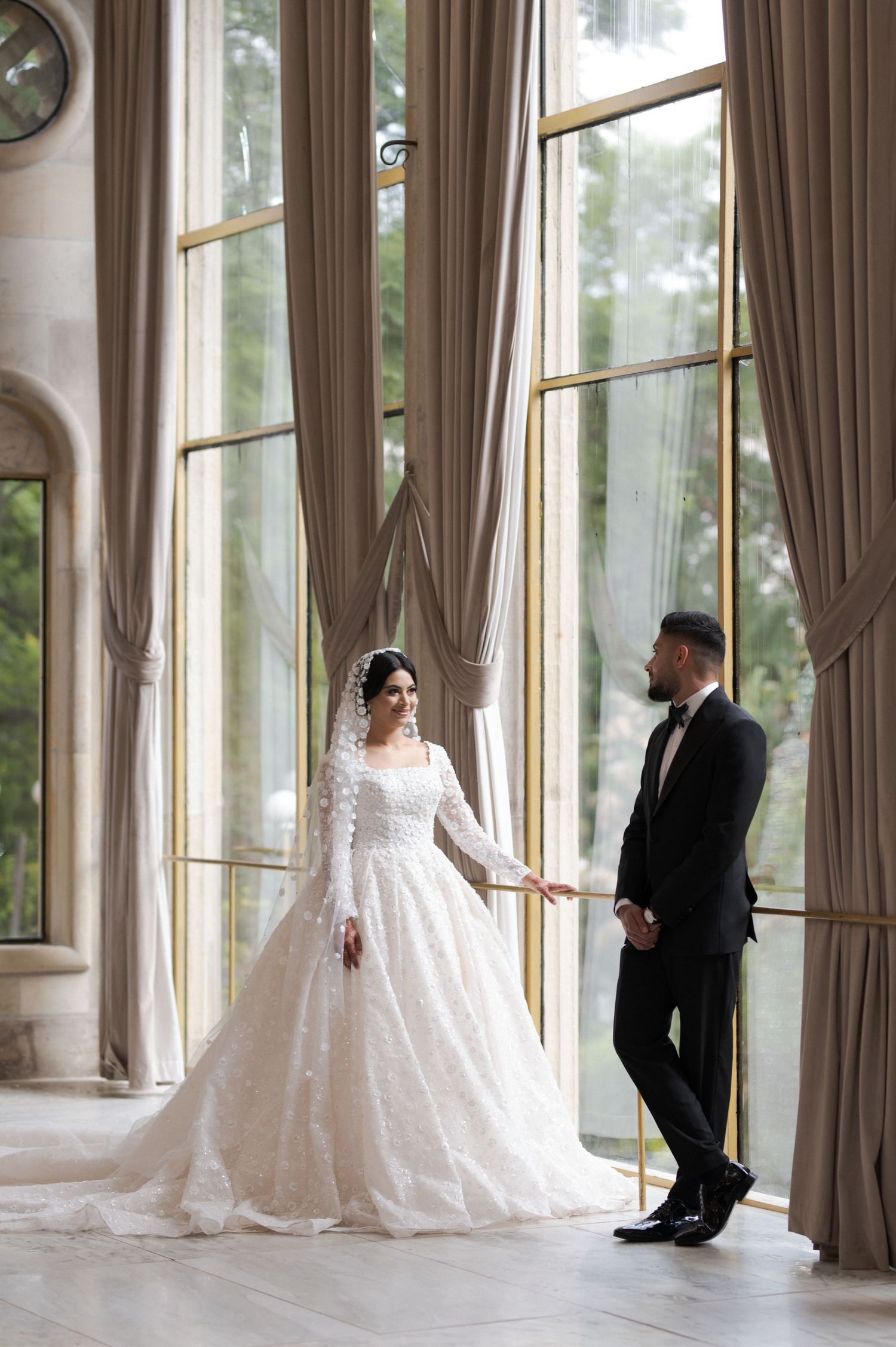 Couple in wedding attire pose by tall windows with flowing curtains in elegant interior.