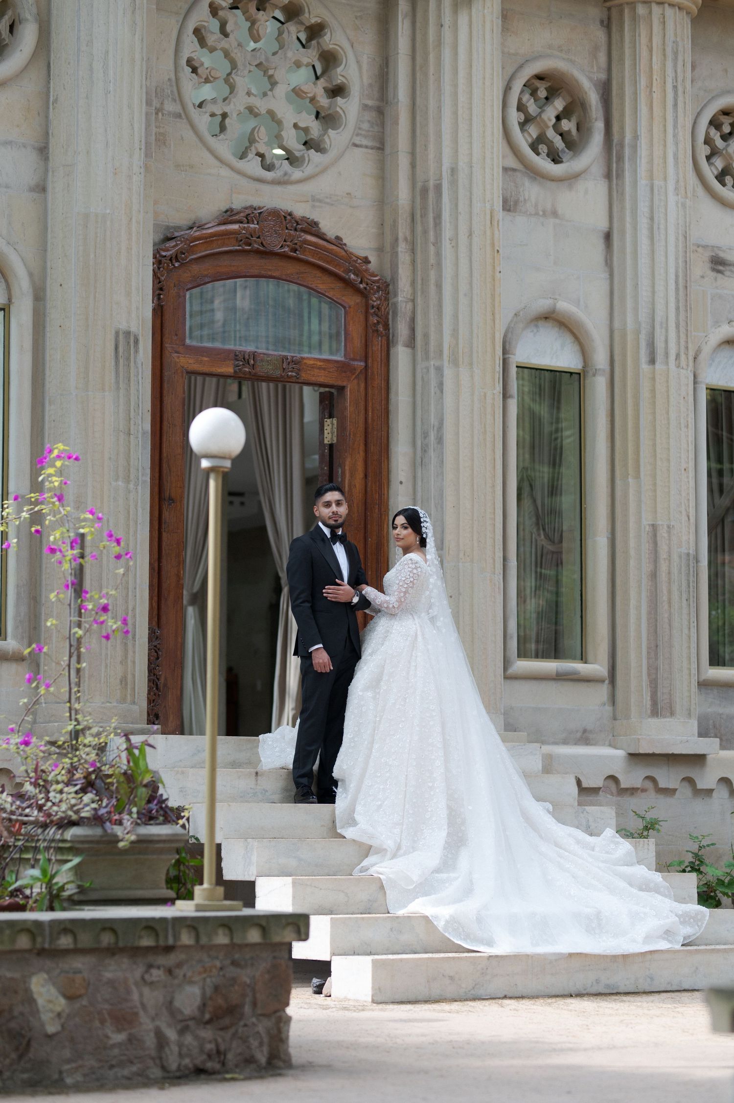A bride and groom stand on elegant stone steps in front of an ornate wooden door at a grand venue.