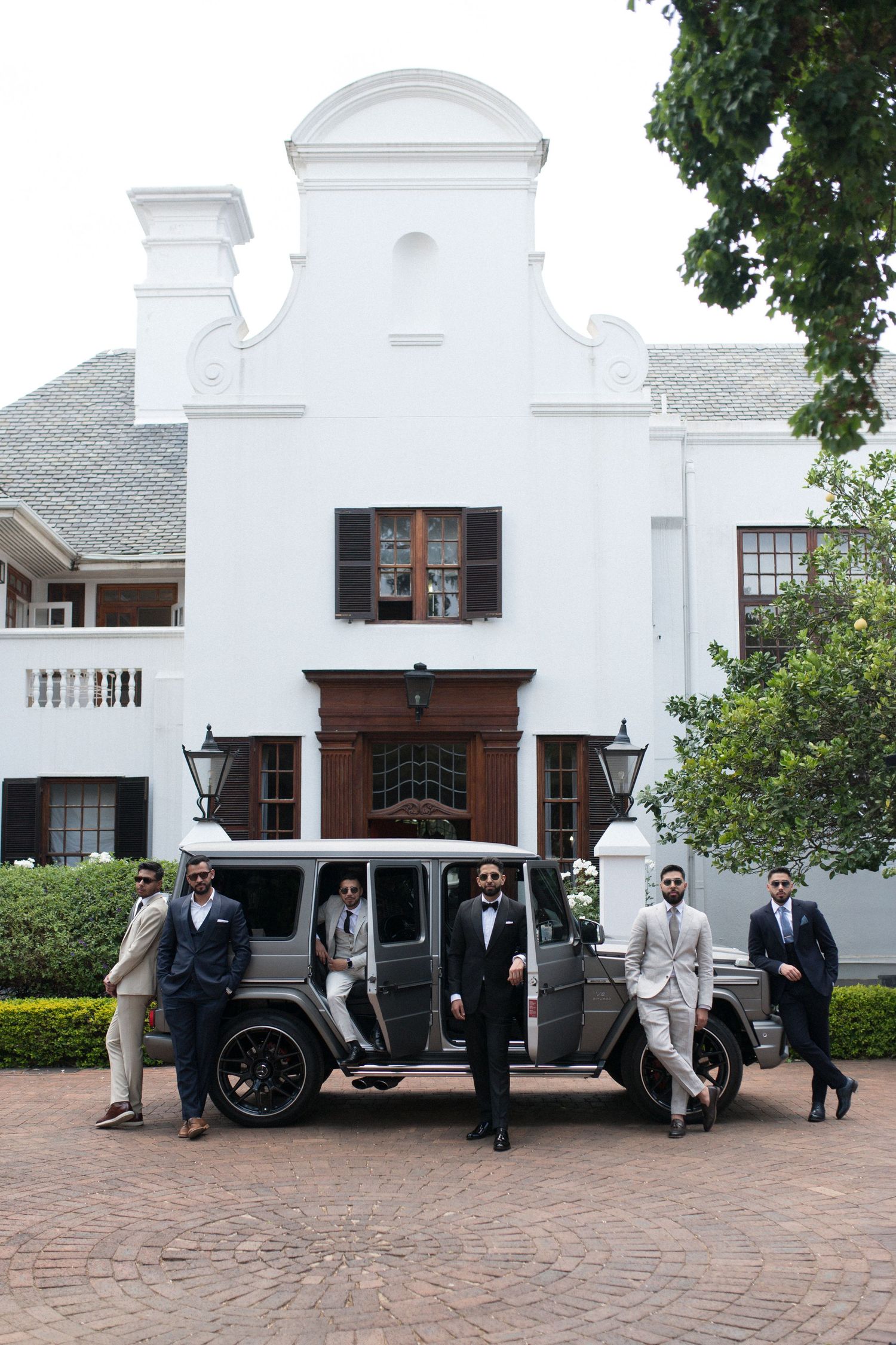 A group standing beside a grey Mercedes G-Wagon in front of a white Cape Dutch style building.