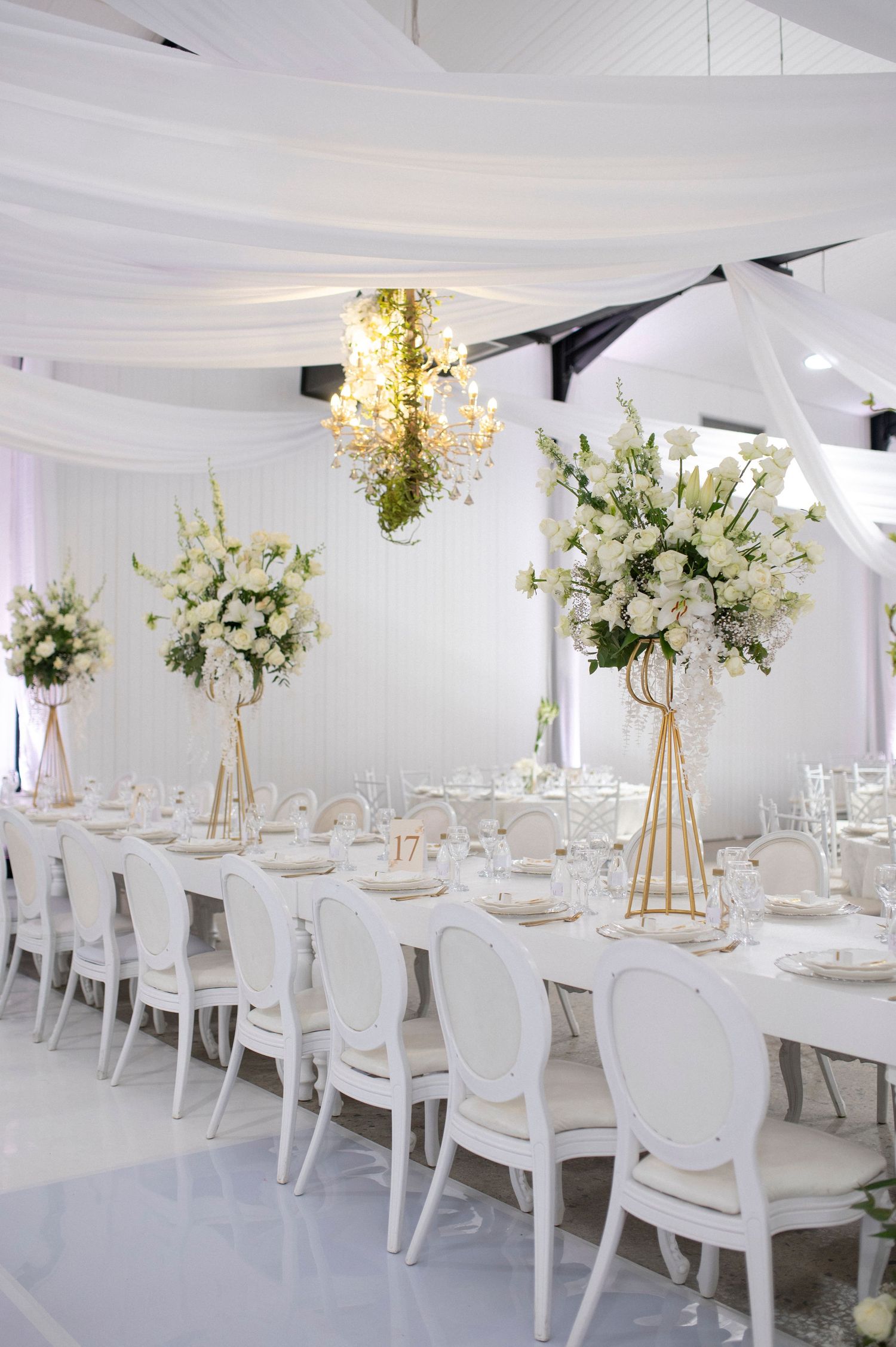 Elegant white wedding reception setup with tall floral centerpieces and luxurious draping.