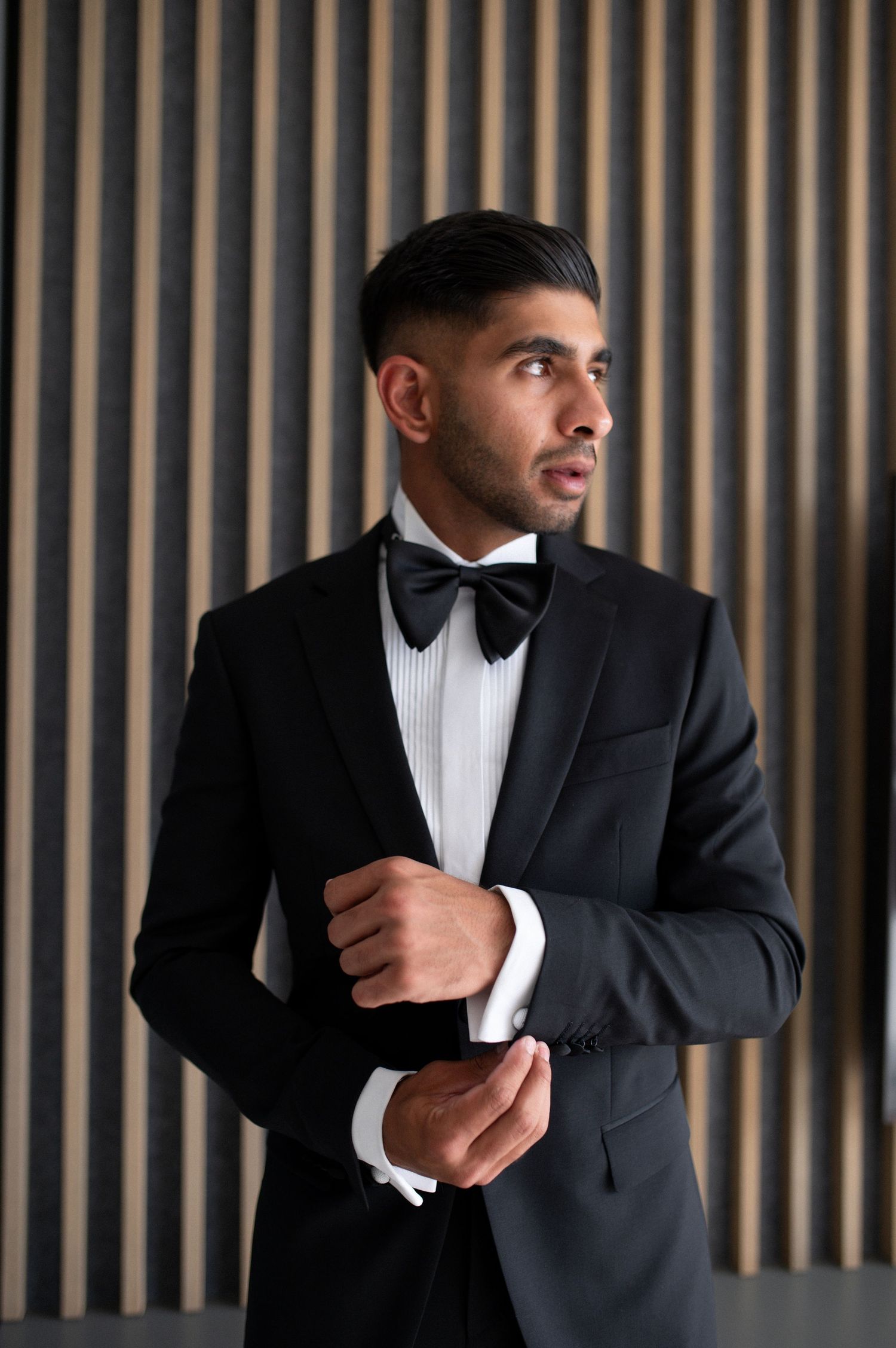 Dapper groom in formal black tuxedo poses against modern striped backdrop.