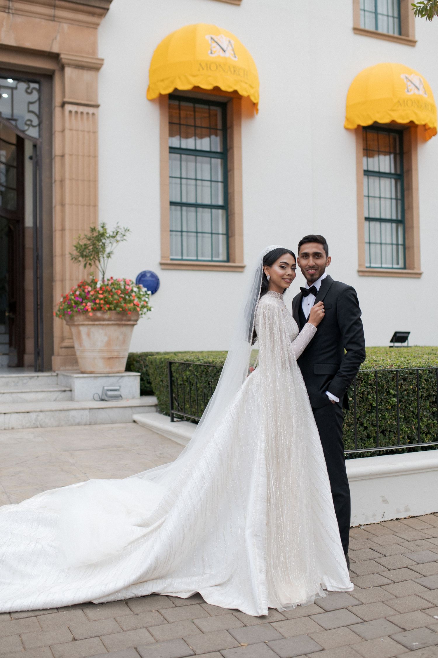 A wedding couple poses together outside a classical building with yellow awnings and manicured hedges.