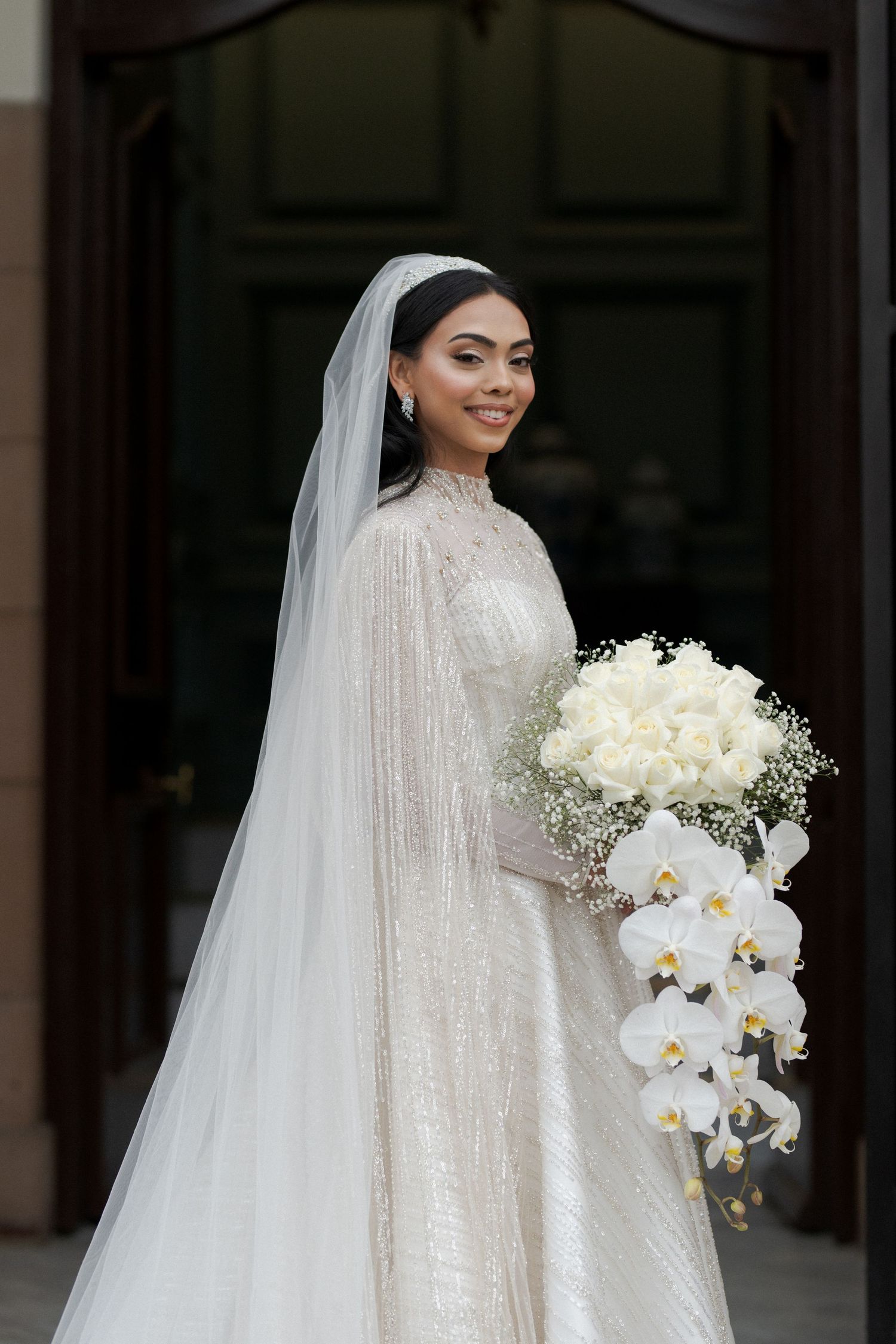 A bride in a long-sleeved sequined wedding gown holds an elegant cascading bouquet of white orchids and roses.