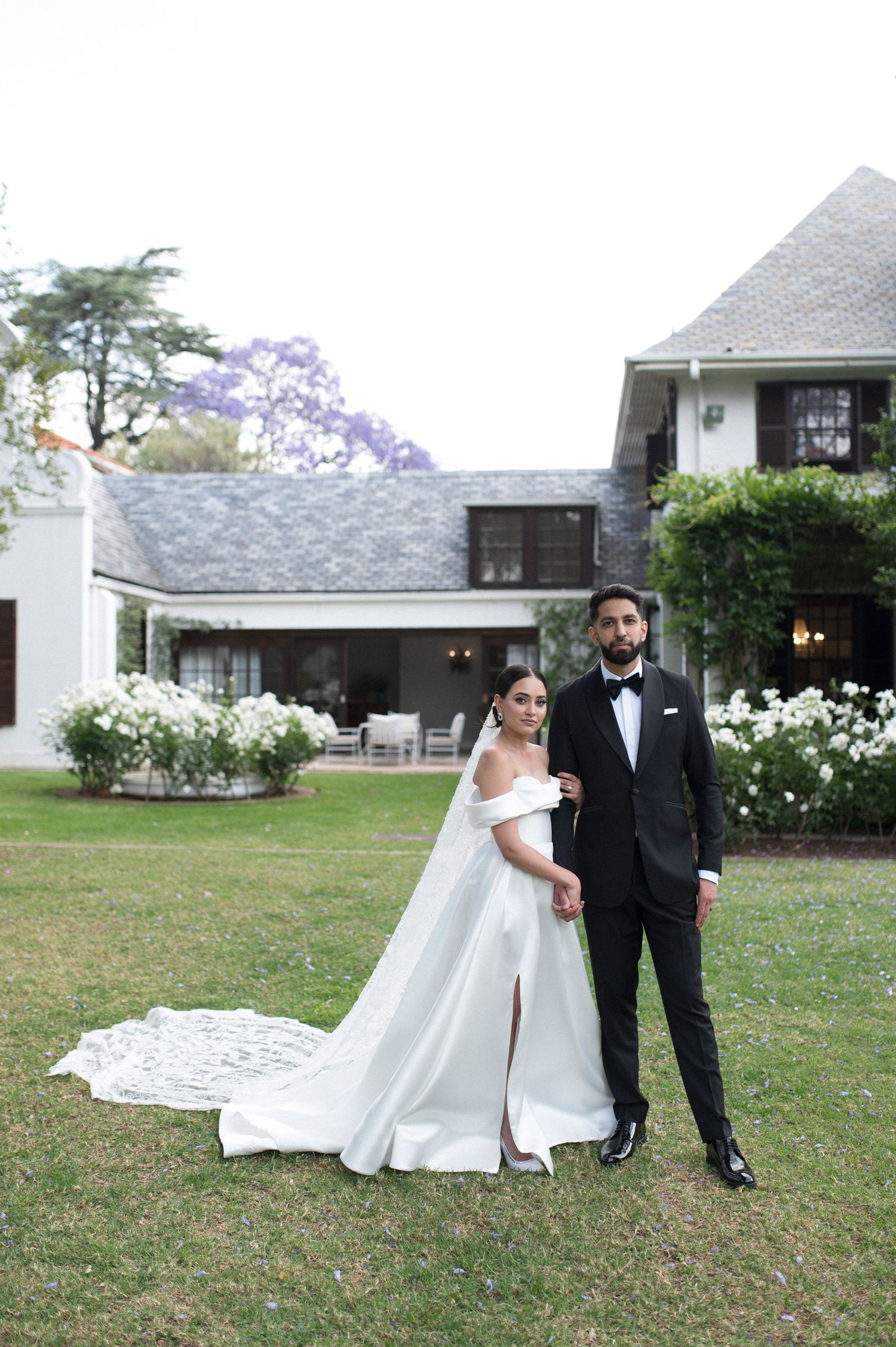 An elegant wedding couple poses on a lawn in front of a beautiful house with white roses and jacaranda trees.