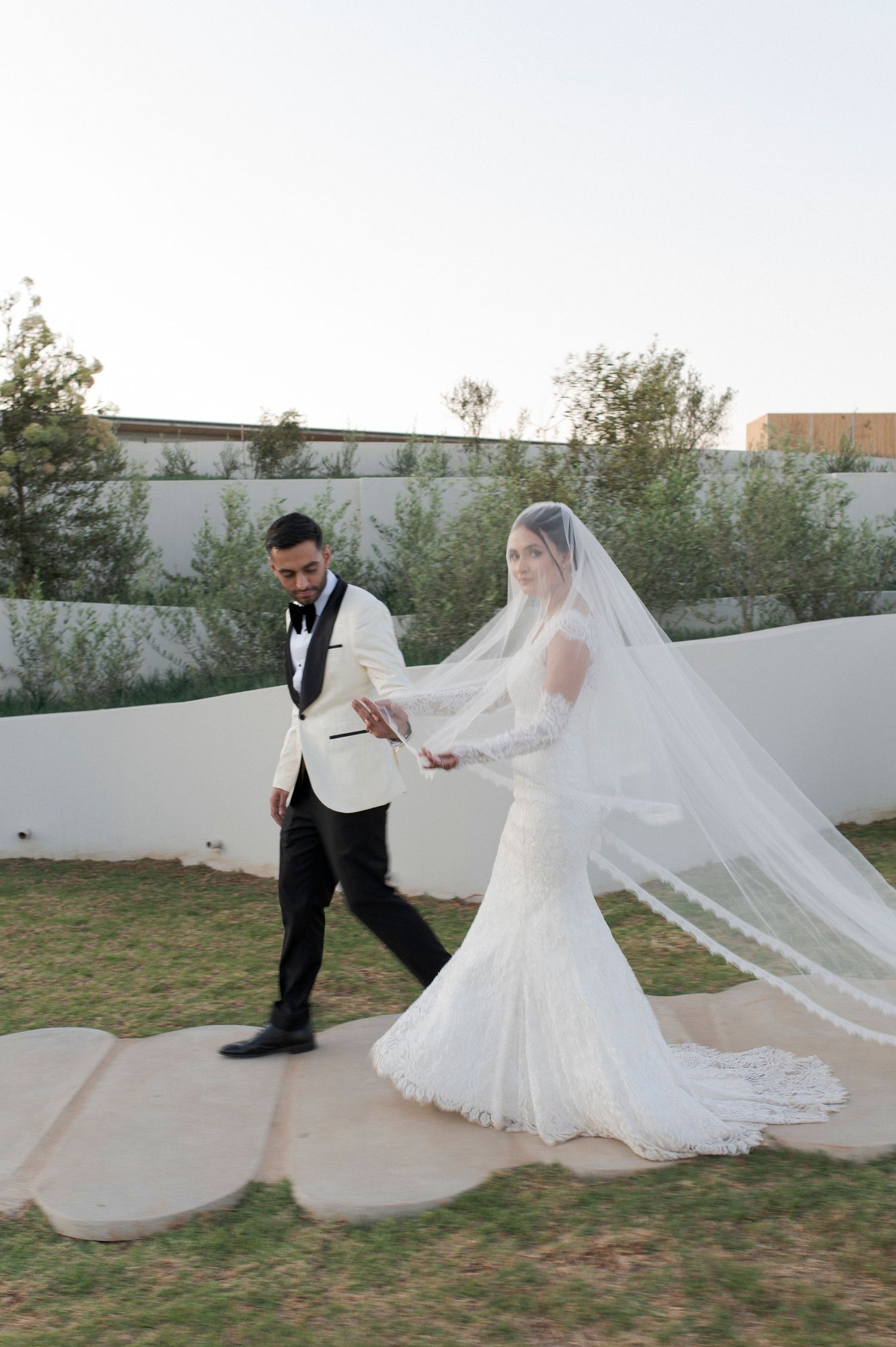 A newlywed couple strolls along a stone pathway in an outdoor garden setting.