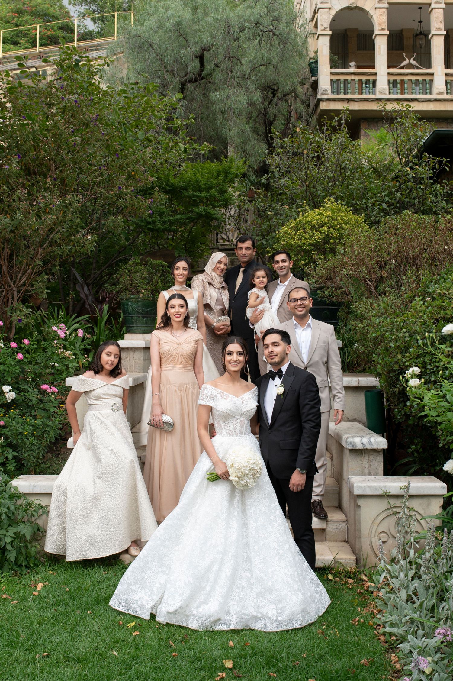 Wedding party poses on stone steps in a lush garden setting with mixed formal attire.