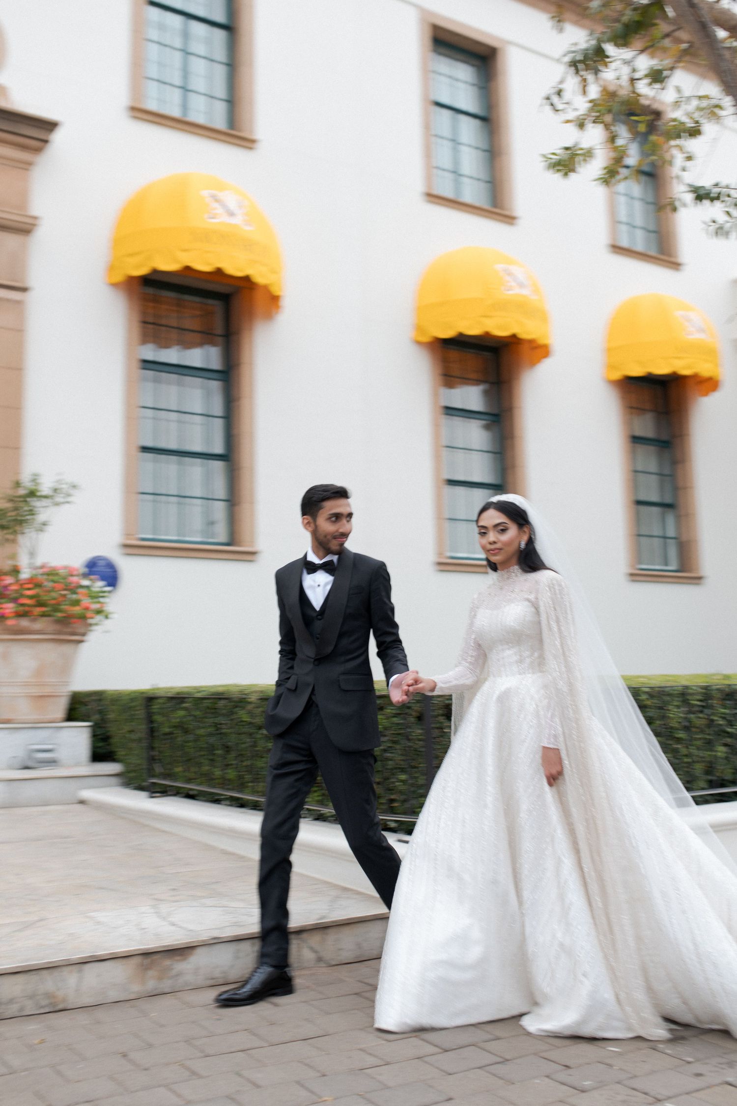An elegant couple walks along a path in front of a building with bright yellow awnings.