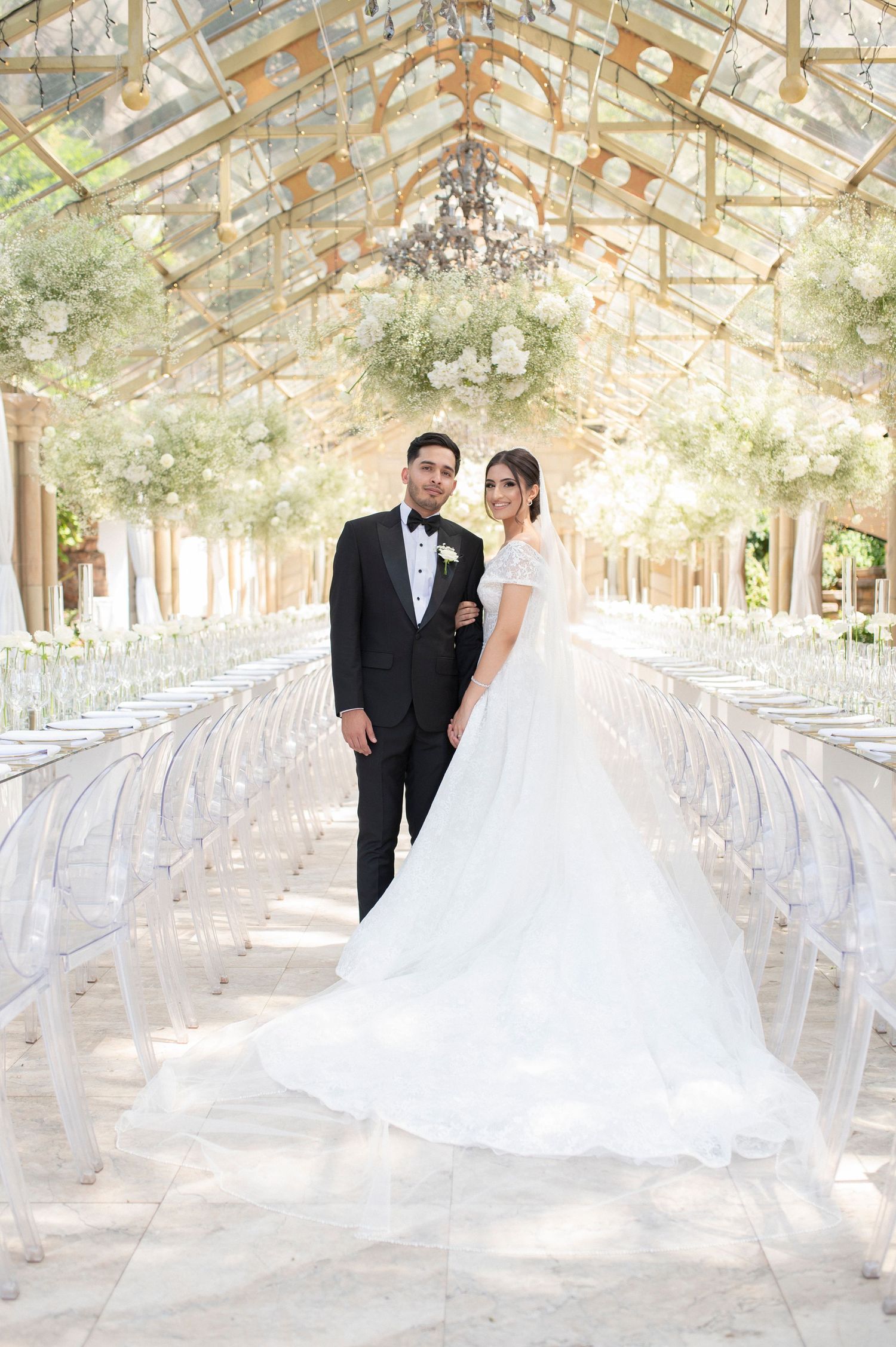 A wedding portrait in a stunning glass-ceiling venue decorated with hanging greenery and ghost chairs.