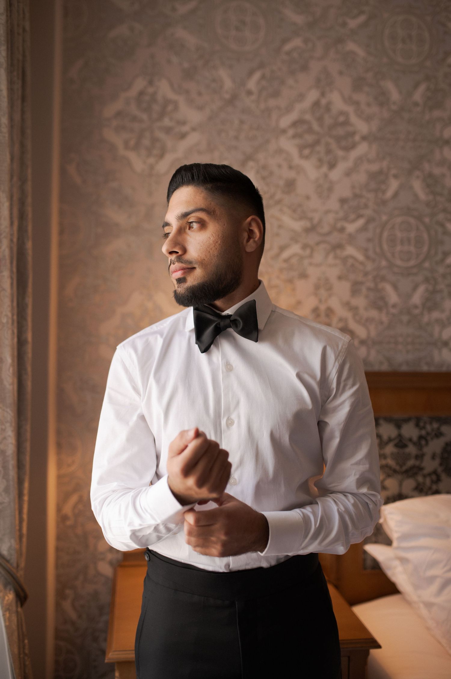 A groom adjusts his cufflinks while wearing a classic white shirt and black bow tie.