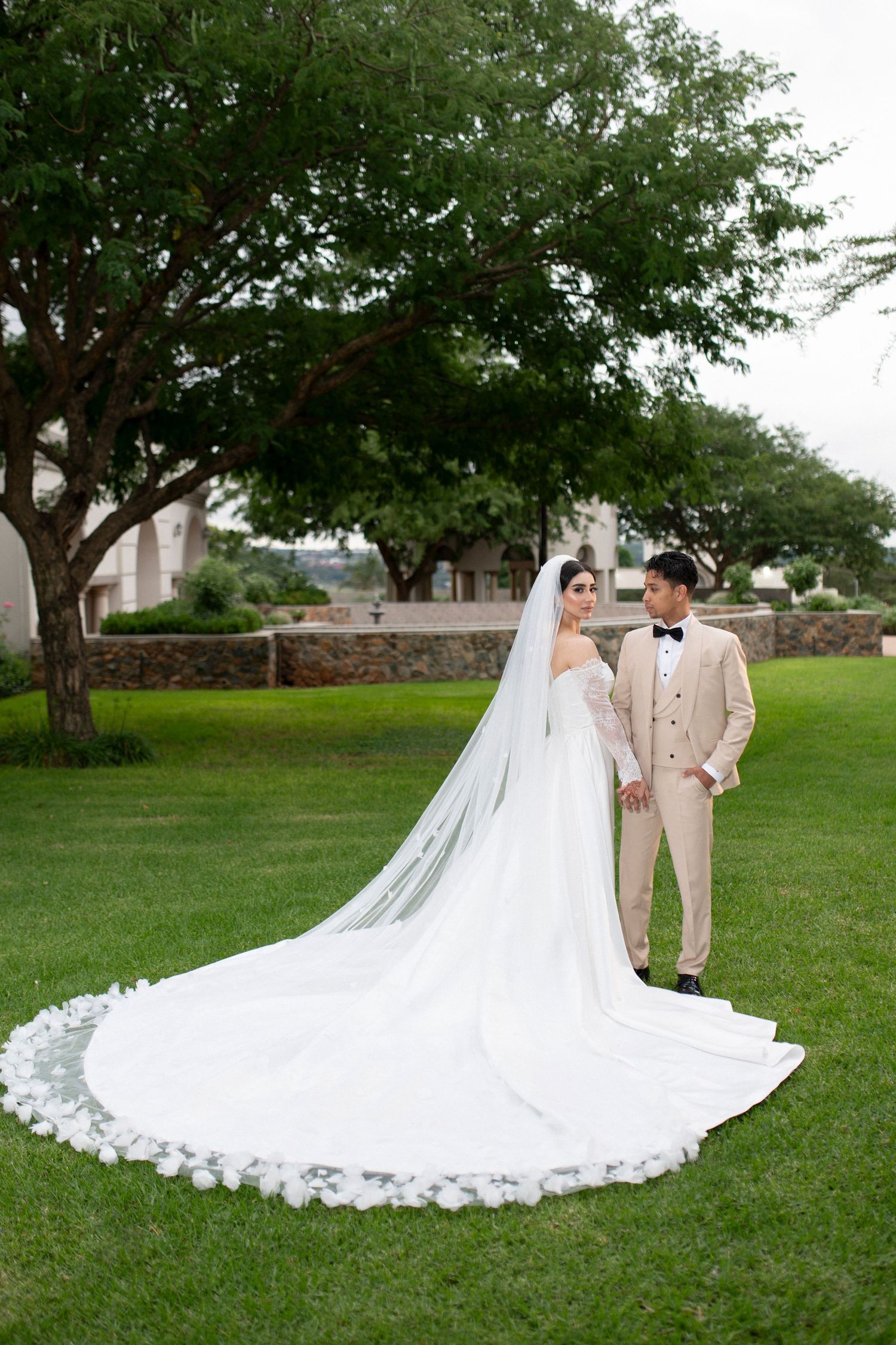 Bride in stunning lace-trimmed cathedral veil poses with groom on manicured lawn.