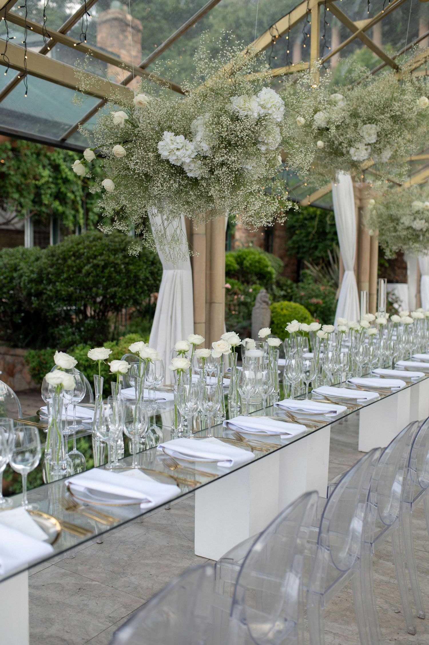 Elegant wedding table setting with crystal chairs and white floral centerpieces in a glass-ceiling venue.