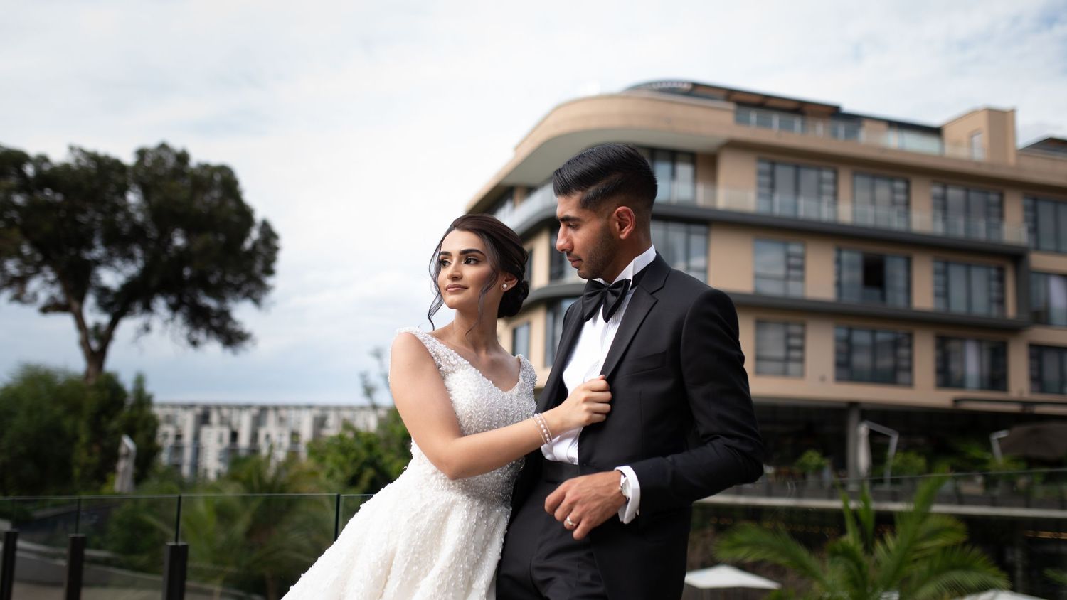 A wedding couple embraces on a balcony with a modern building in the background.