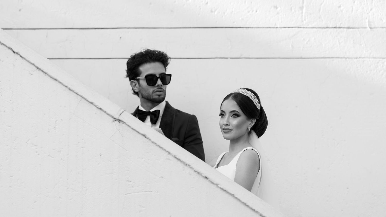 A couple in formal attire poses on stairs, with the groom in sunglasses and bride wearing a headband.