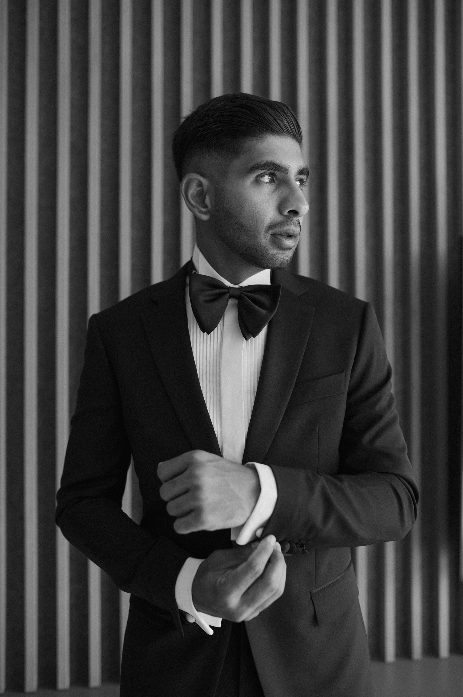 Groom in classic black tuxedo and bow tie adjusting cufflinks against striped wall.