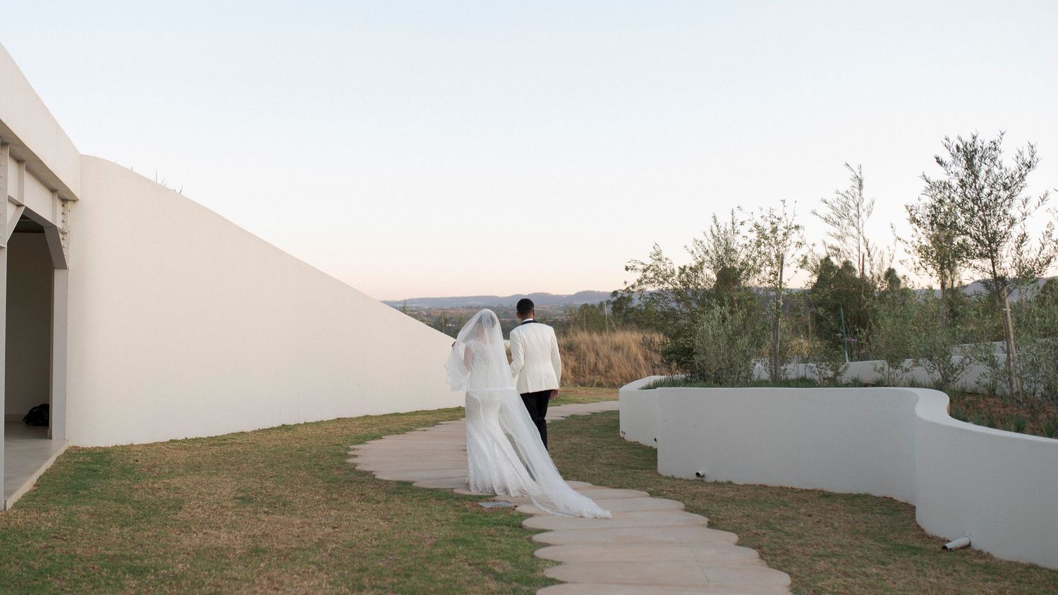 A wedding couple walks along a winding path between white modern architectural walls at dusk.