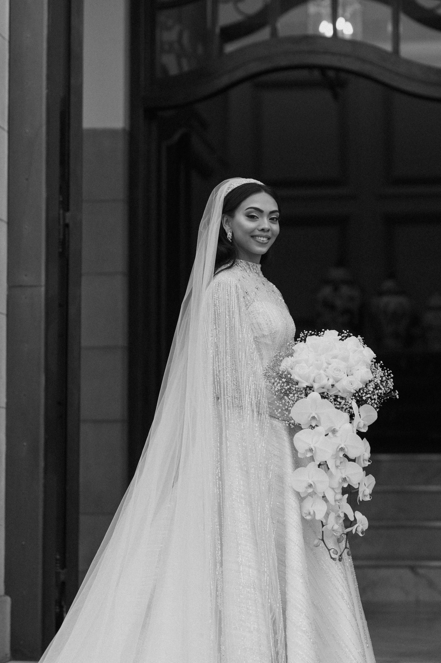 A black and white portrait of a bride holding a cascading bouquet while standing by elegant doors.