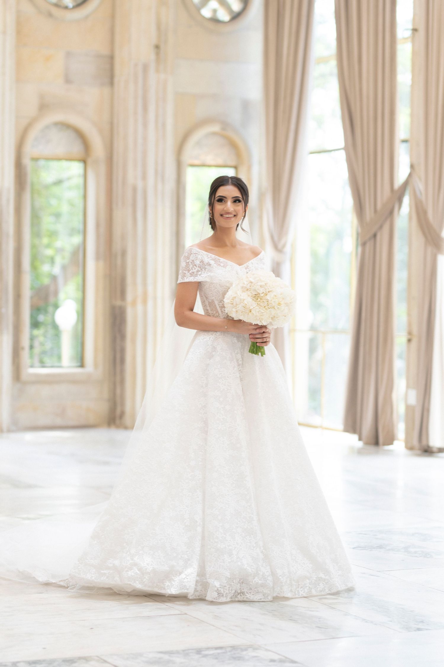 A full-length portrait of a bride in an off-shoulder wedding gown holding a white bouquet in an ornate room.