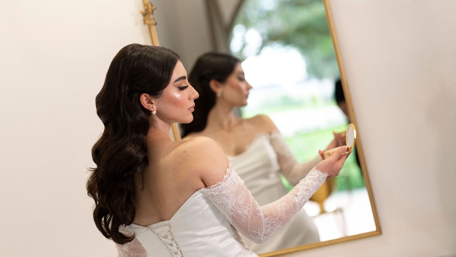 Bride in white lace gown looks in mirror while getting ready with classic wavy hair.