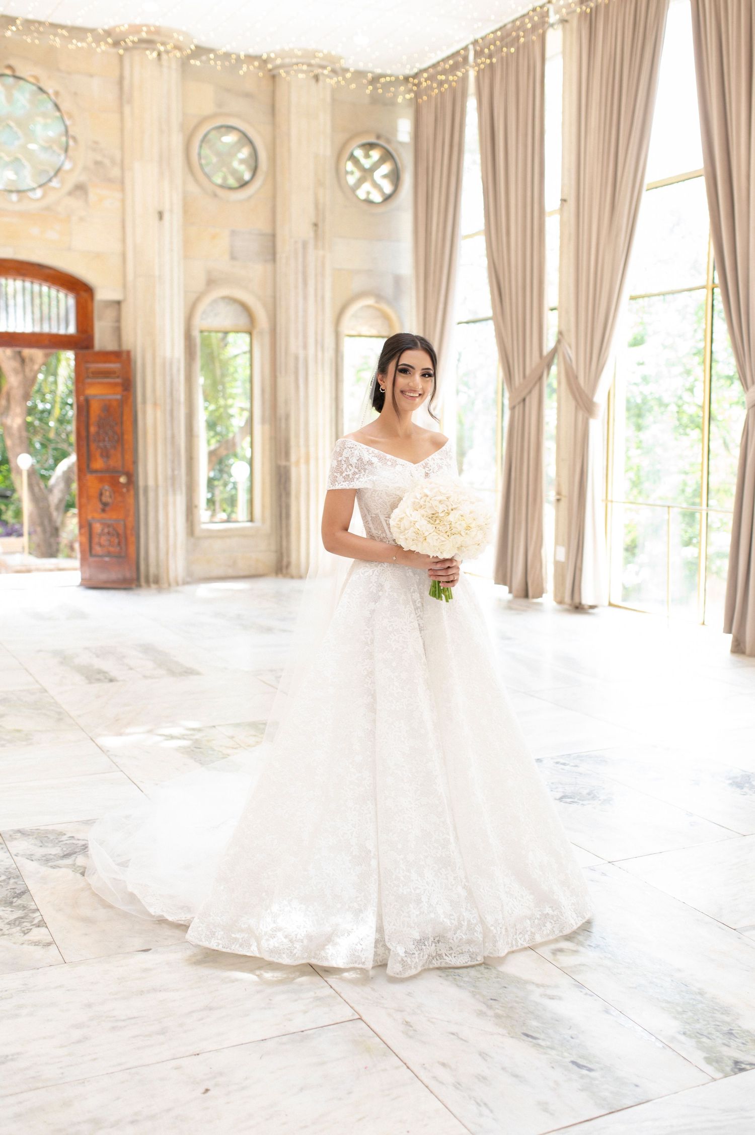 A bride poses in a lace wedding gown inside an ornate room with tall windows and marble floors.