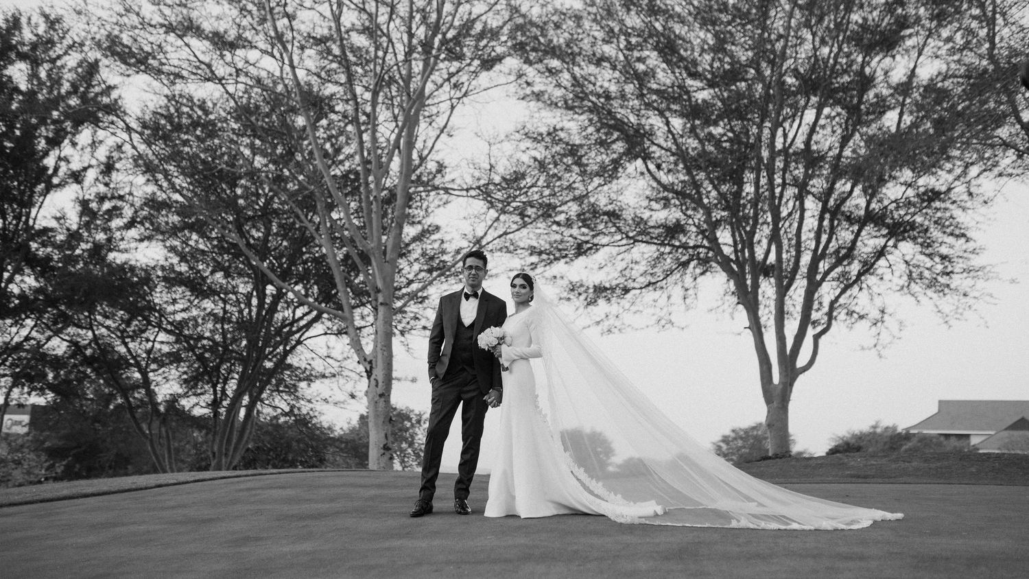 Dramatic black and white photo of newlyweds standing beneath trees with flowing veil.