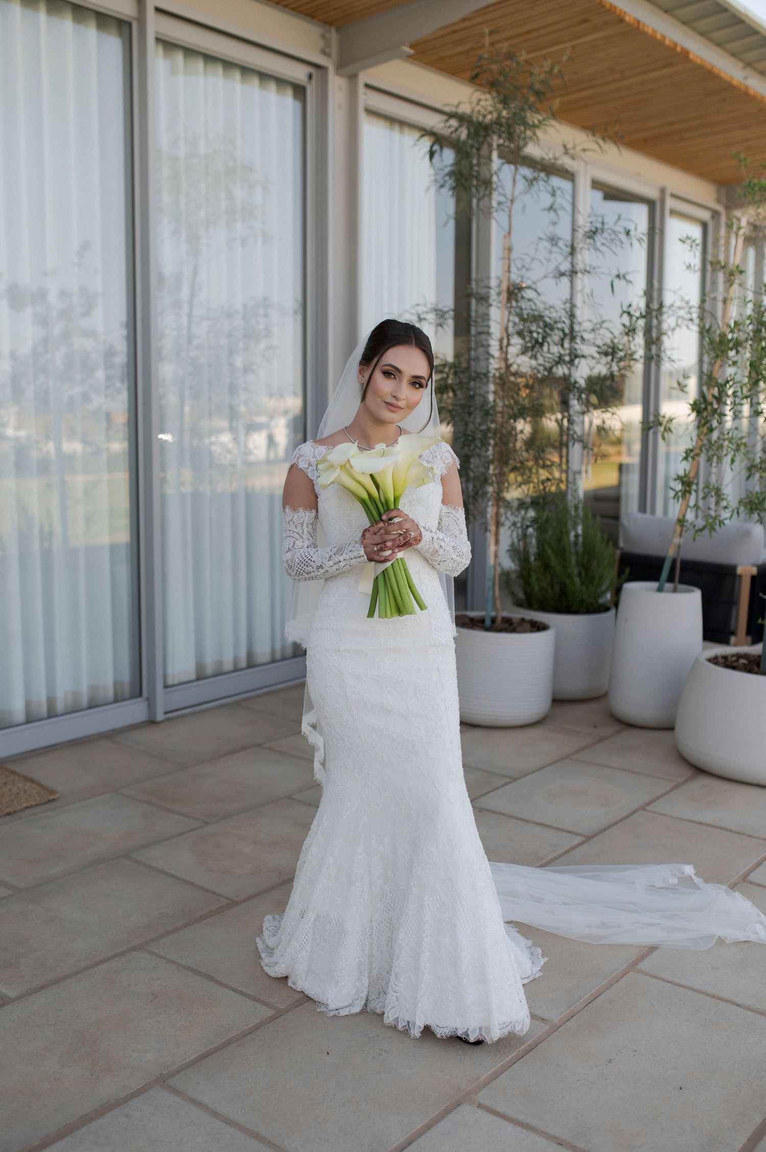 Bride in elegant lace wedding dress holding white calla lily bouquet on outdoor terrace with potted plants.