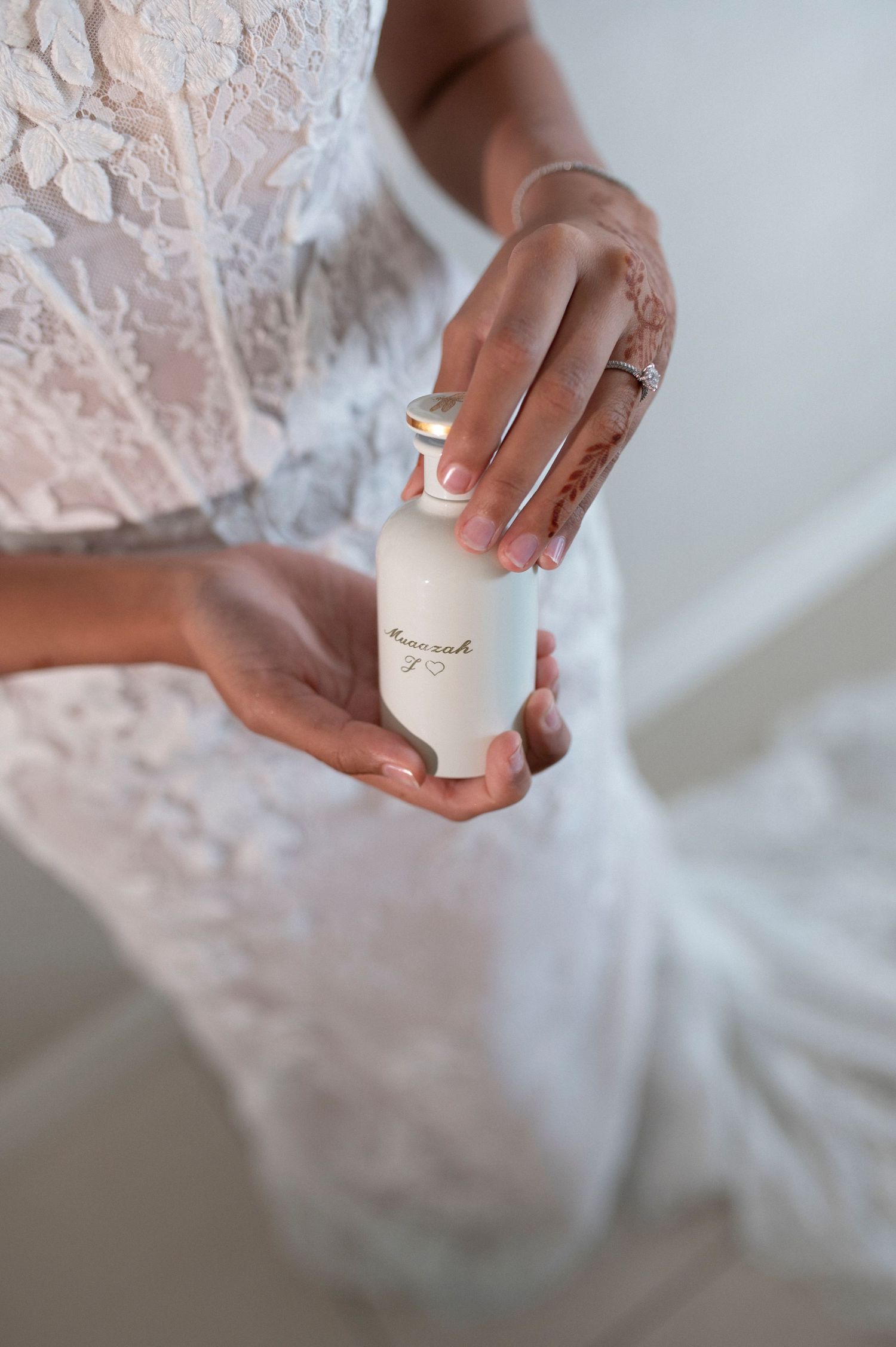 Close-up of hands holding a white perfume or cosmetic bottle against lace fabric in soft lighting.