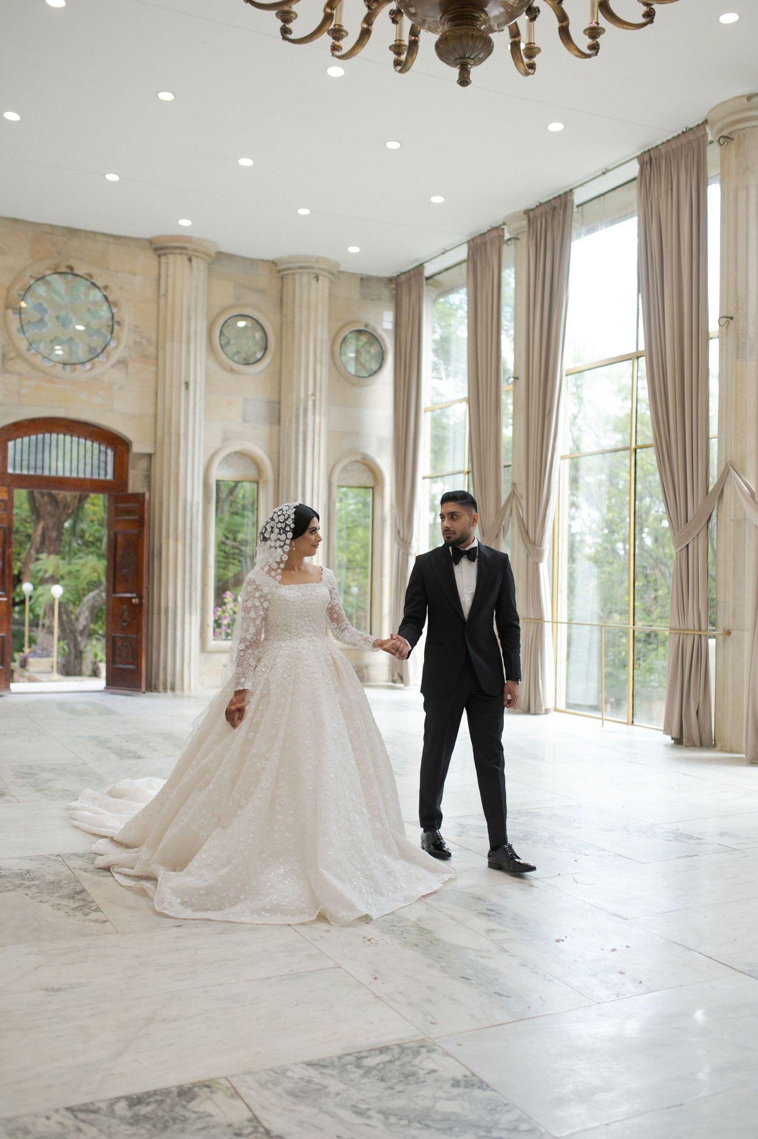 Newlyweds stand in an elegant ballroom with floor-to-ceiling windows and chandelier overhead.