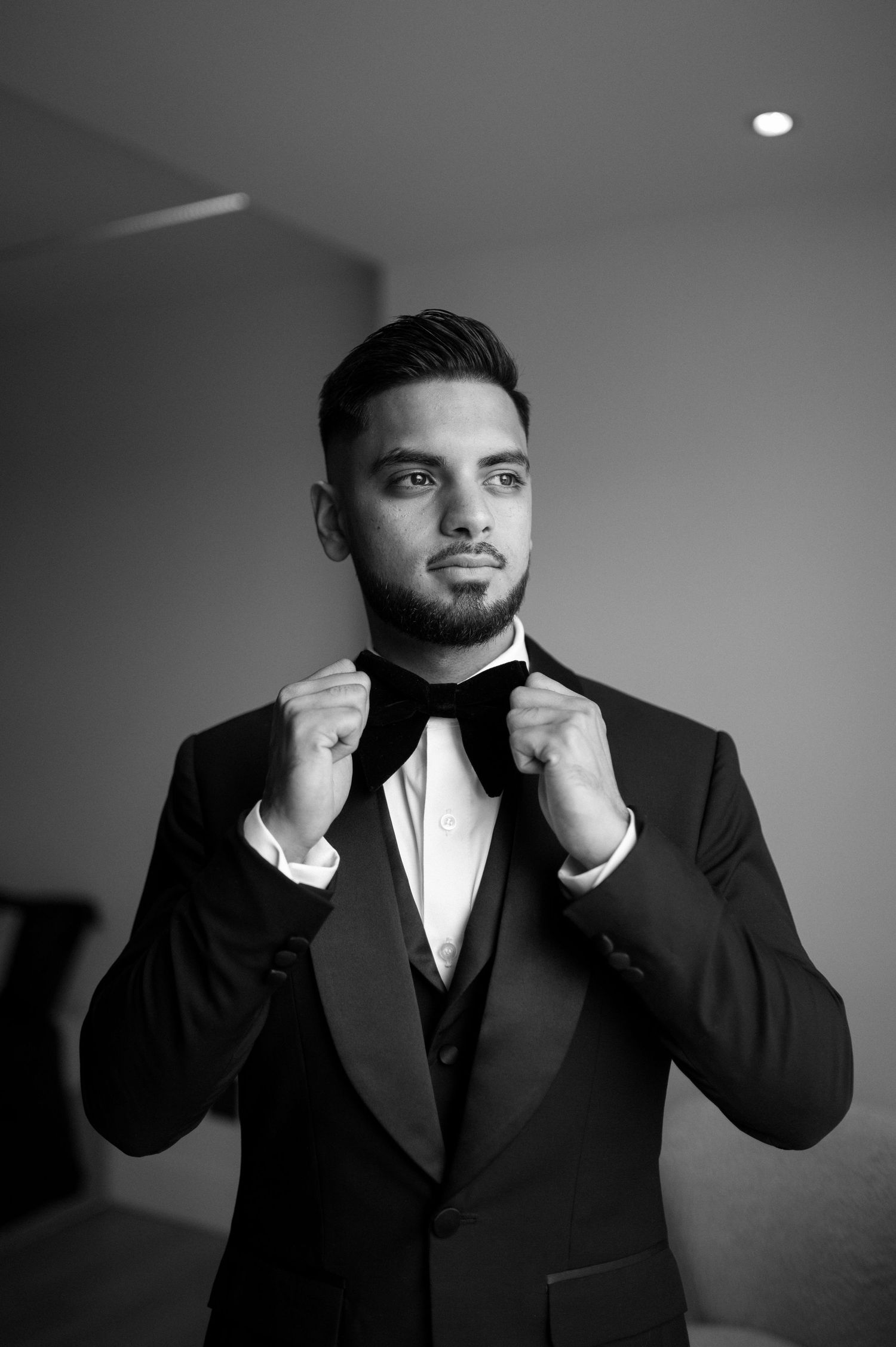 Formal portrait of groom adjusting bowtie in classic black tuxedo.