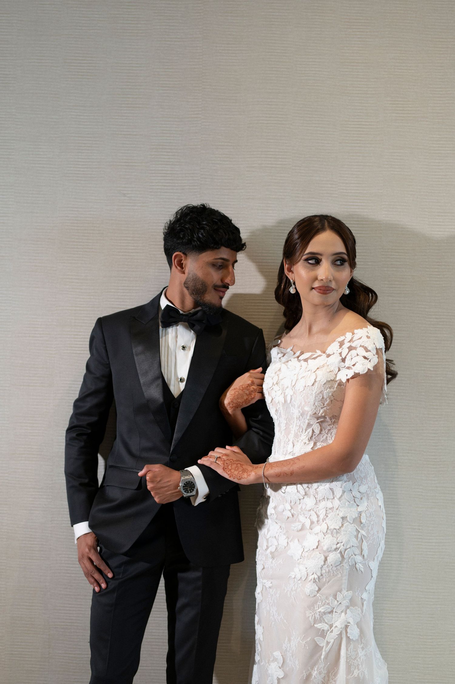 A formal couple in wedding attire posing together against a neutral wall.