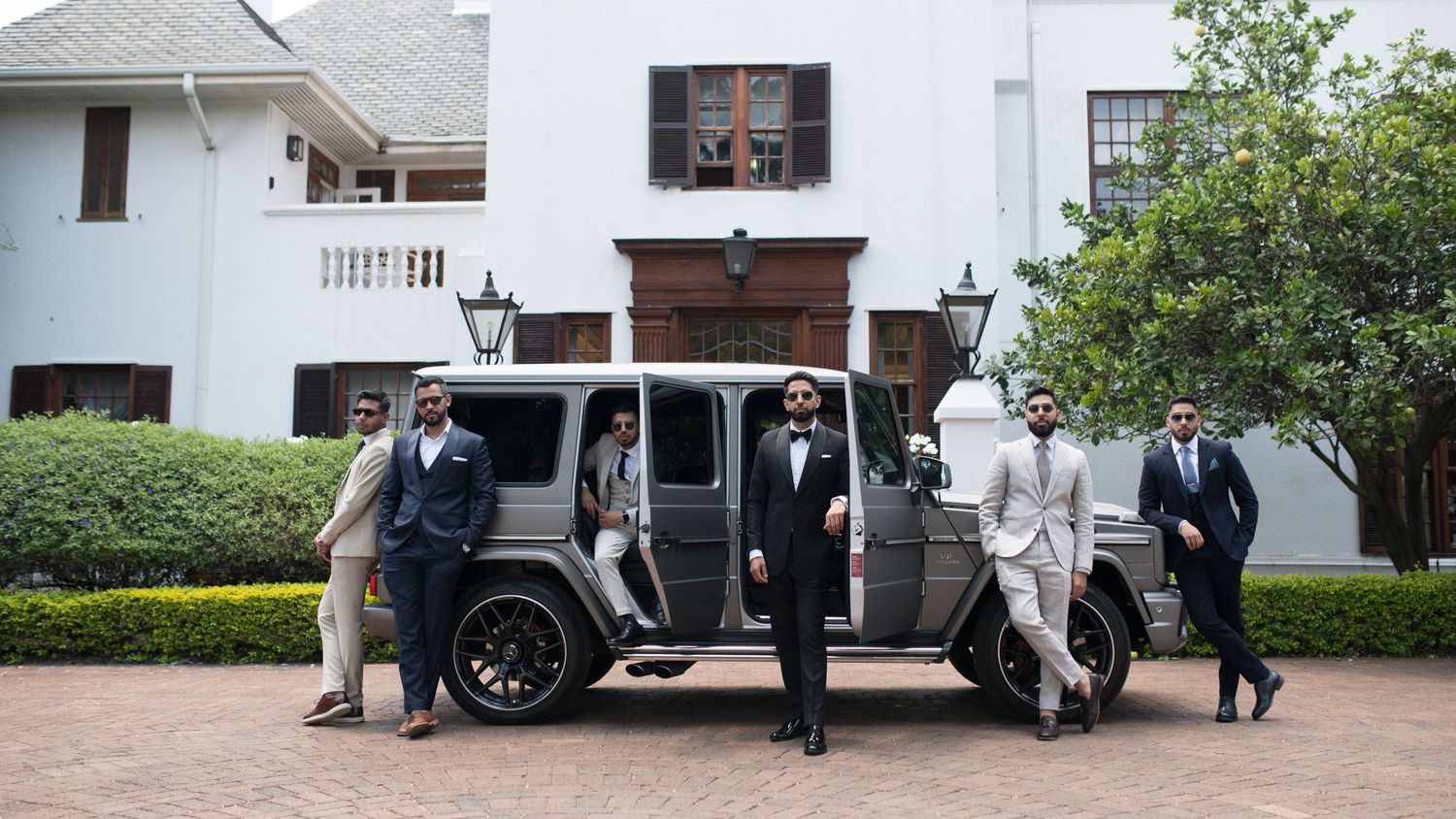 Well-dressed wedding party poses with a grey Mercedes G-Wagon outside a colonial mansion.