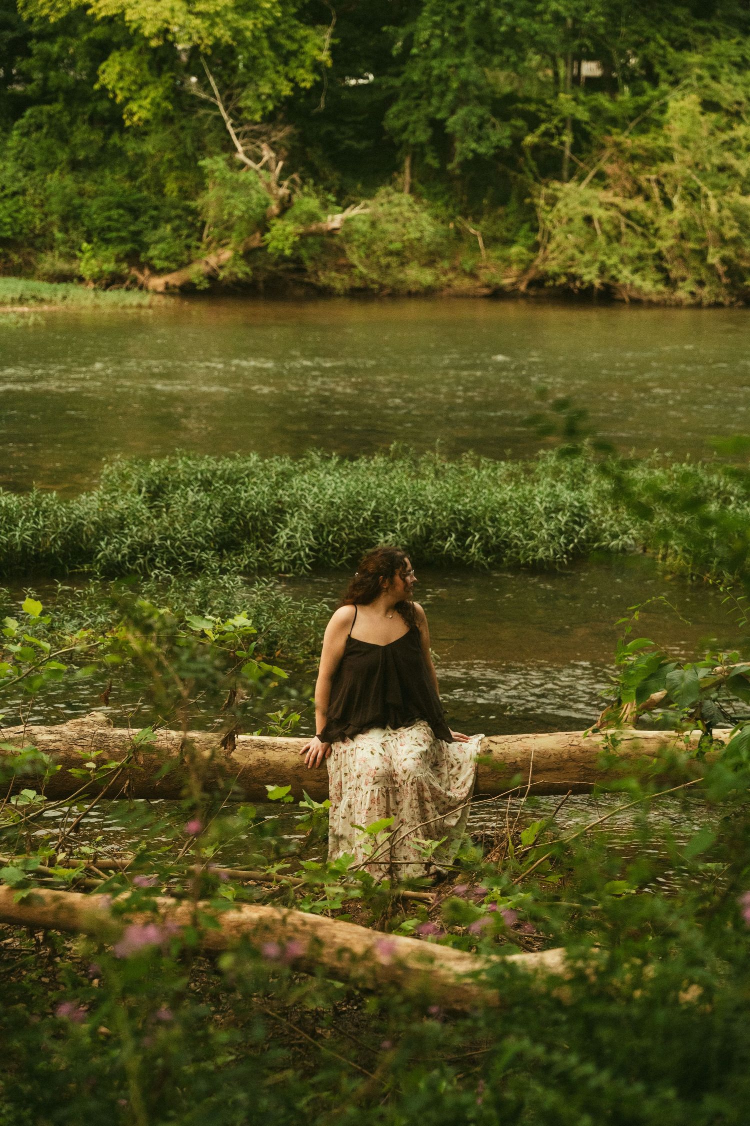 Woman in floral skirt sits on fallen log by a serene river surrounded by lush green forest.