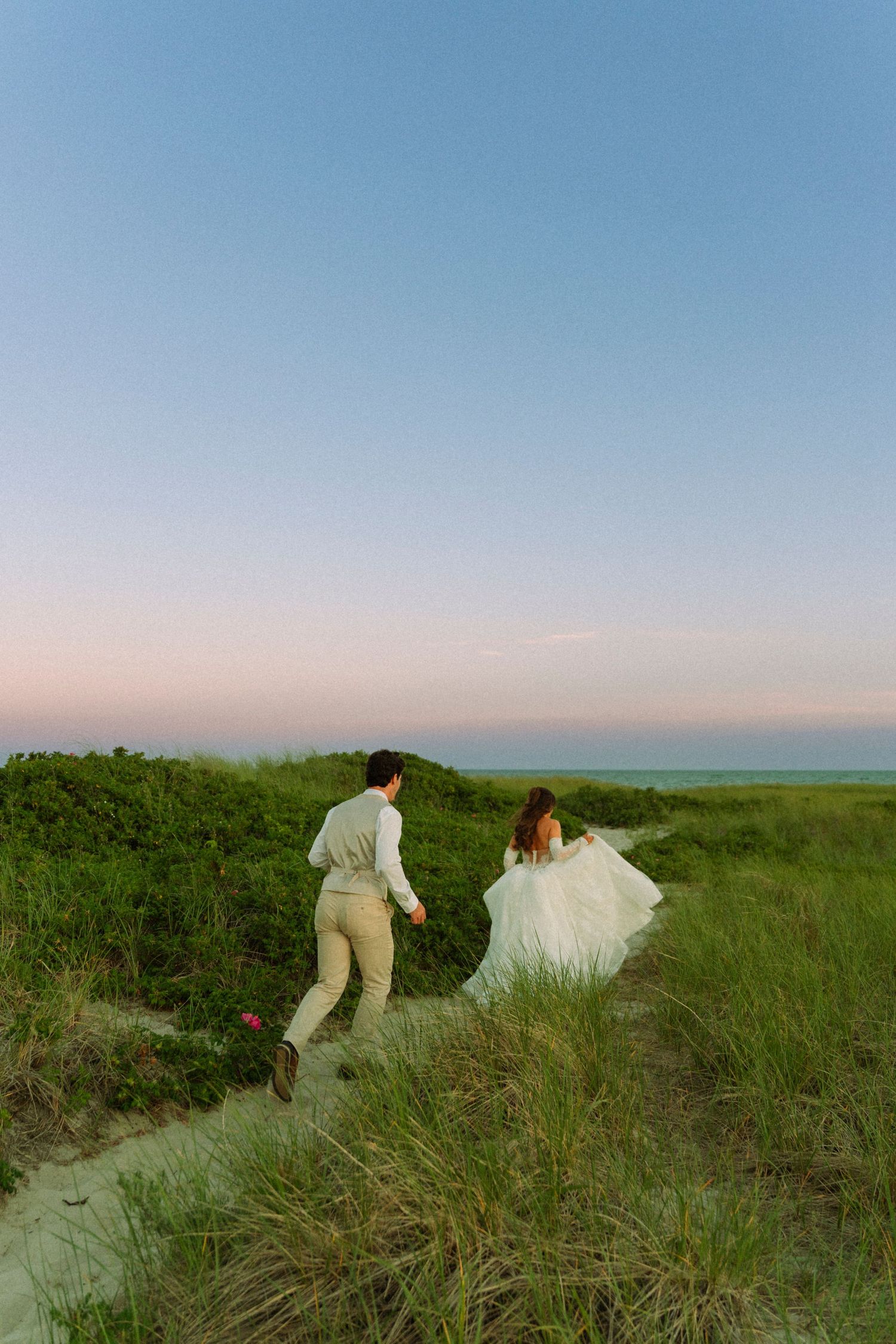 Bride and groom running through green coastal dunes at sunset, bride's white gown flowing in the breeze.
