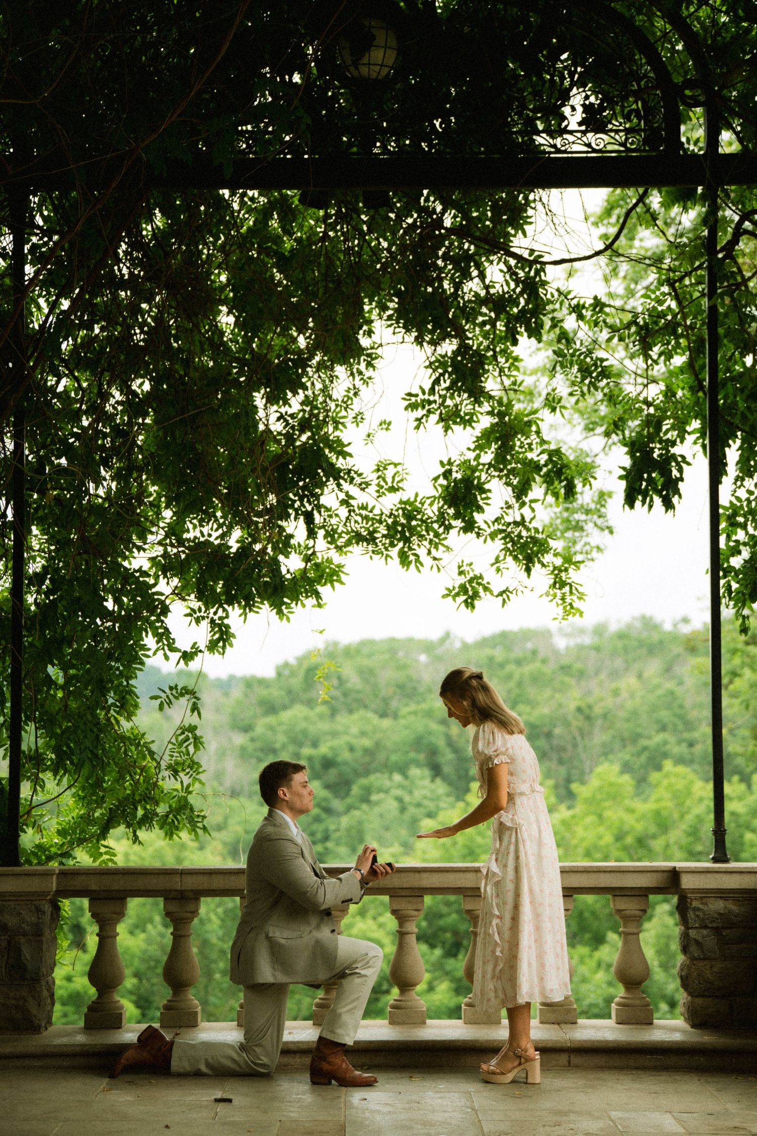 Man proposing on one knee to woman on elegant stone balcony surrounded by lush green trees.
