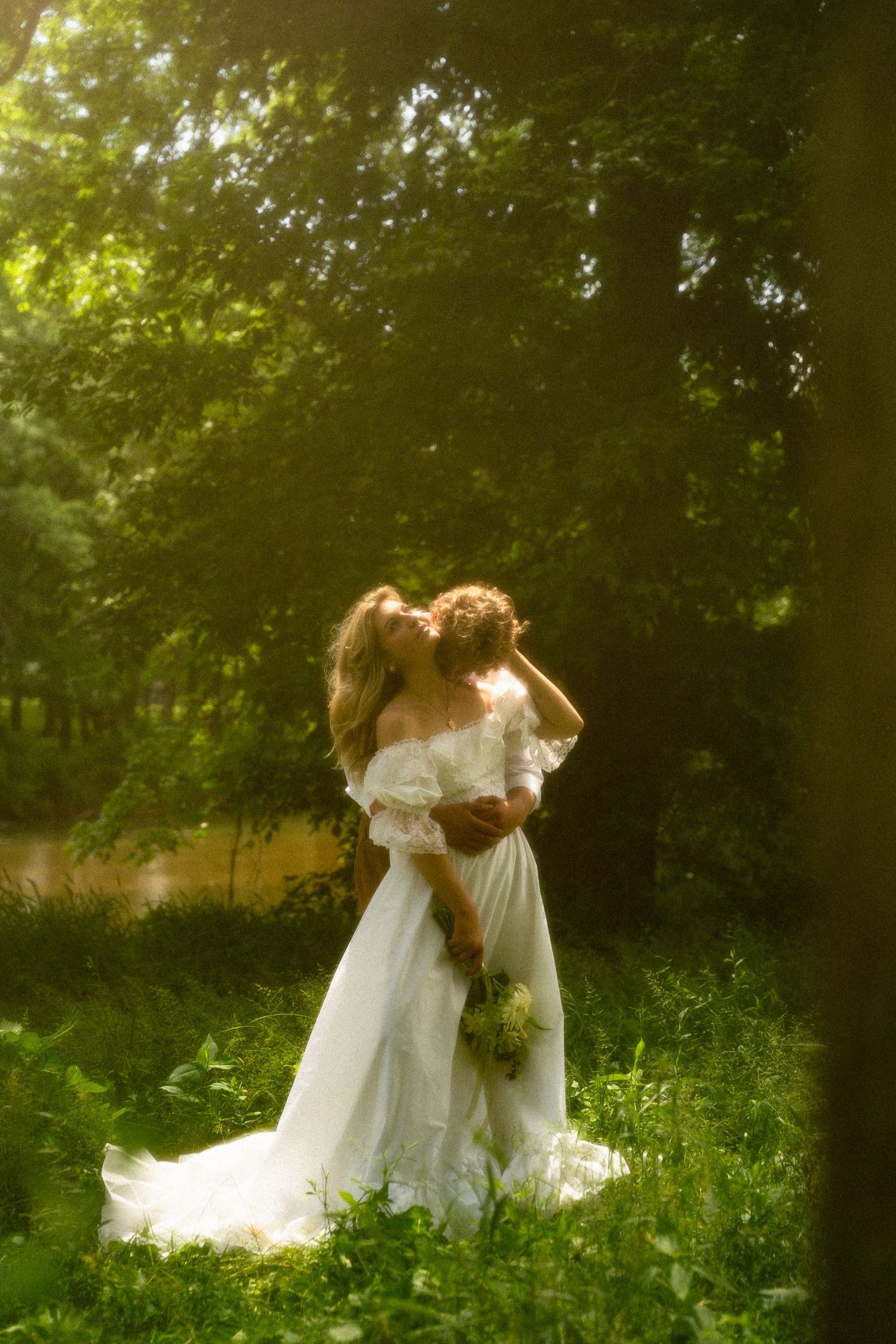 Two people in flowing white dresses embrace in sunlit garden setting.