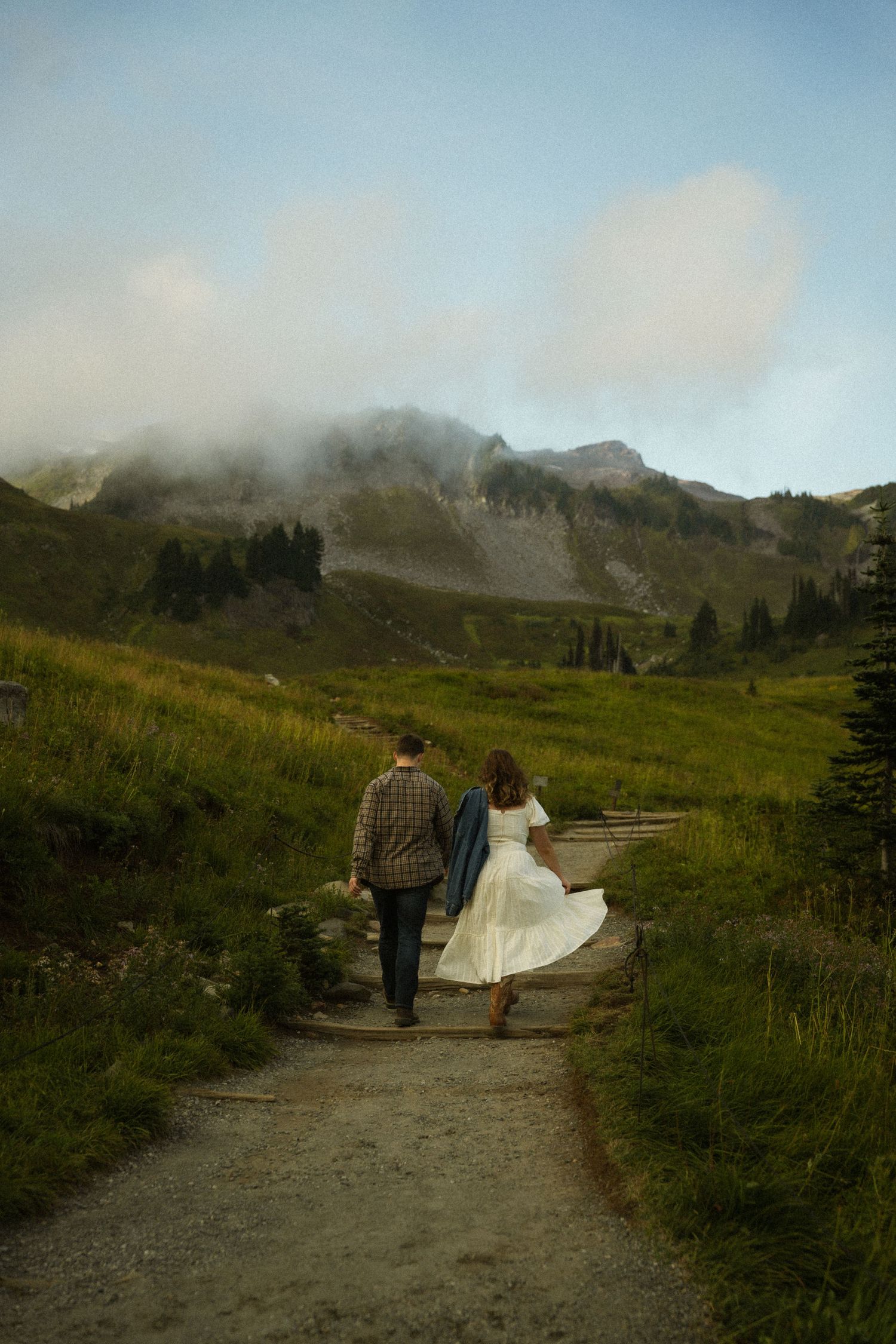 Couple walks down mountain path with foggy alpine scenery and wildflowers in background.