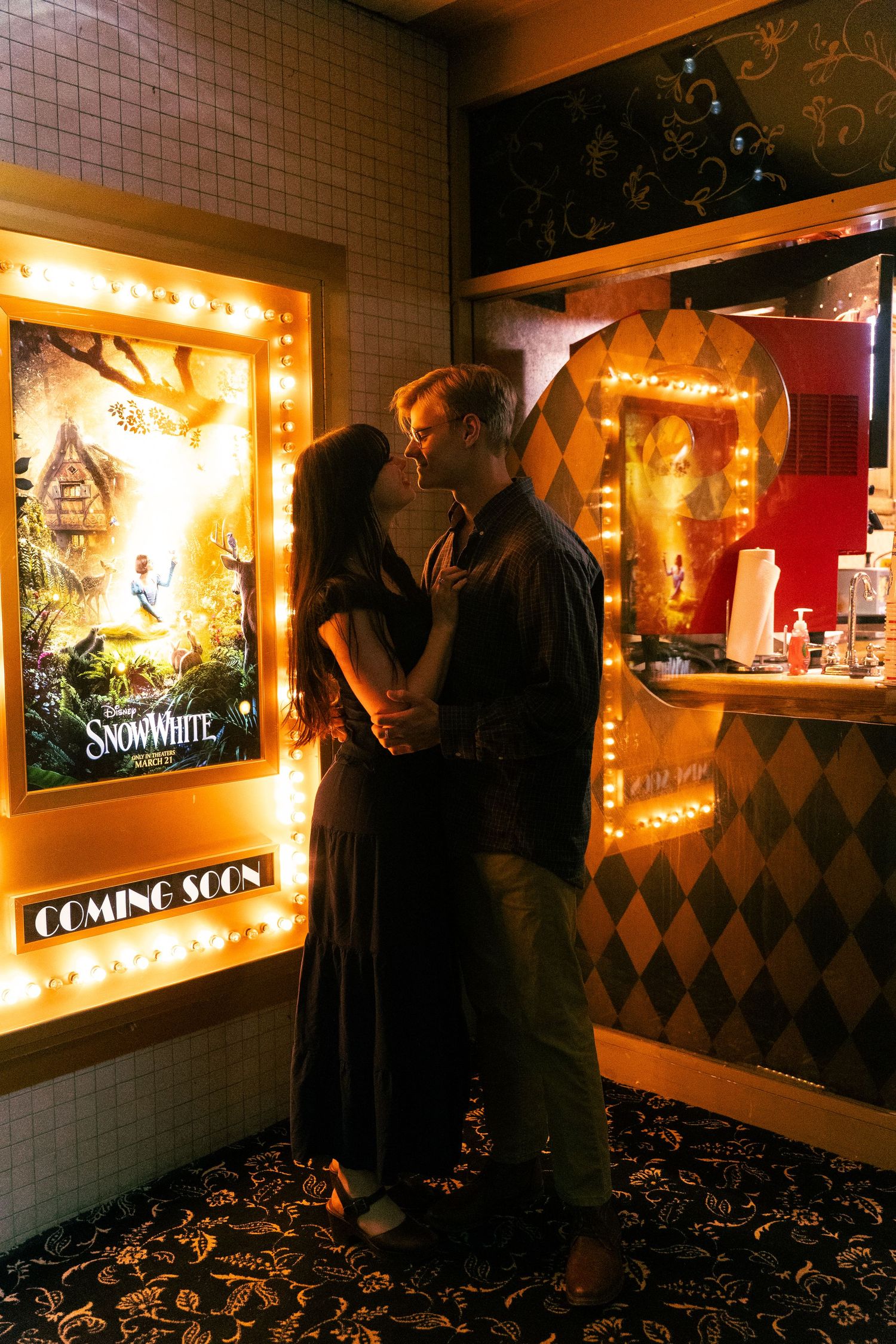 A romantic moment in a dimly lit movie theater lobby with vintage-style illuminated posters and decorative lighting.