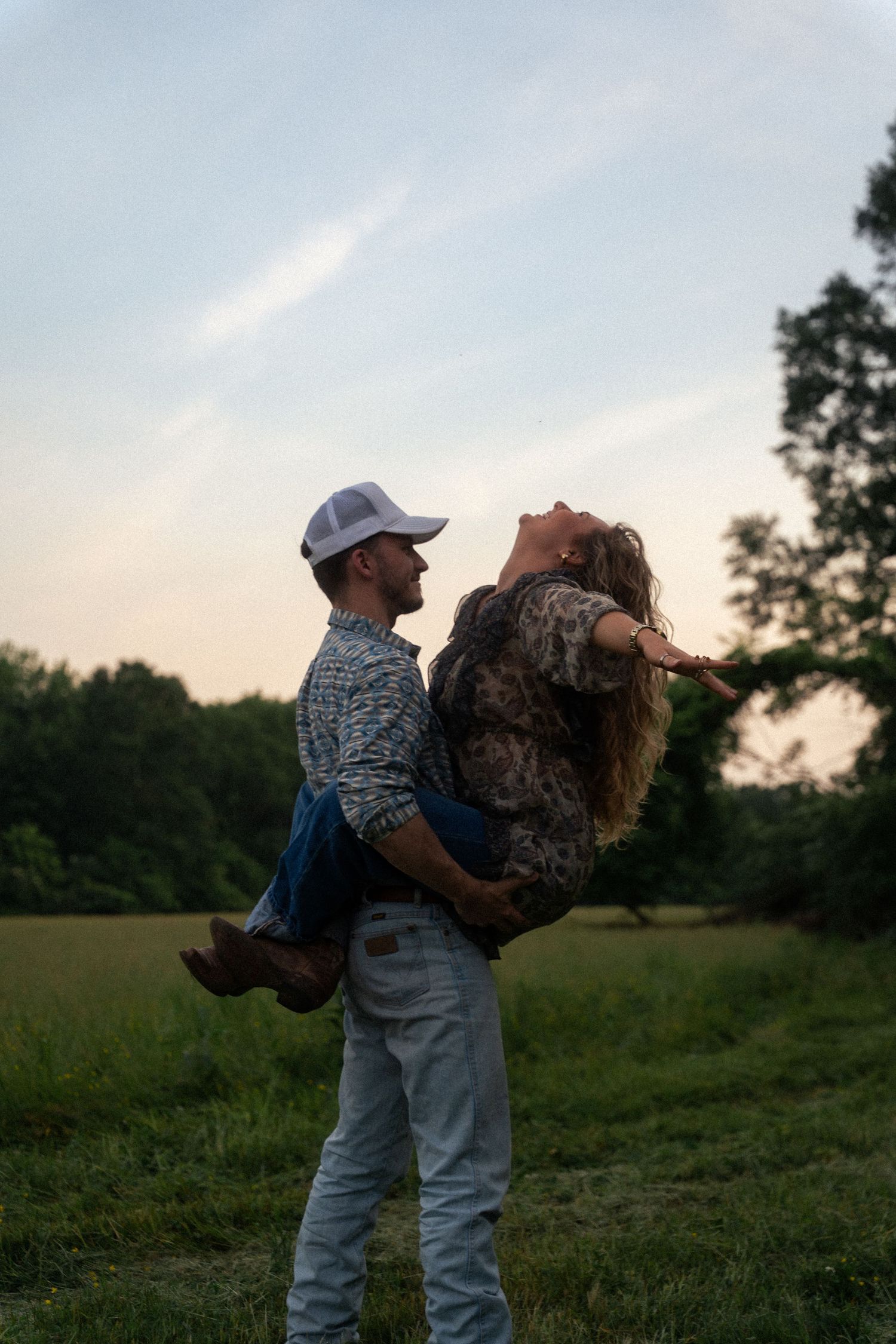 A couple shares a playful moment in a grassy field at sunset, with one person lifting the other.