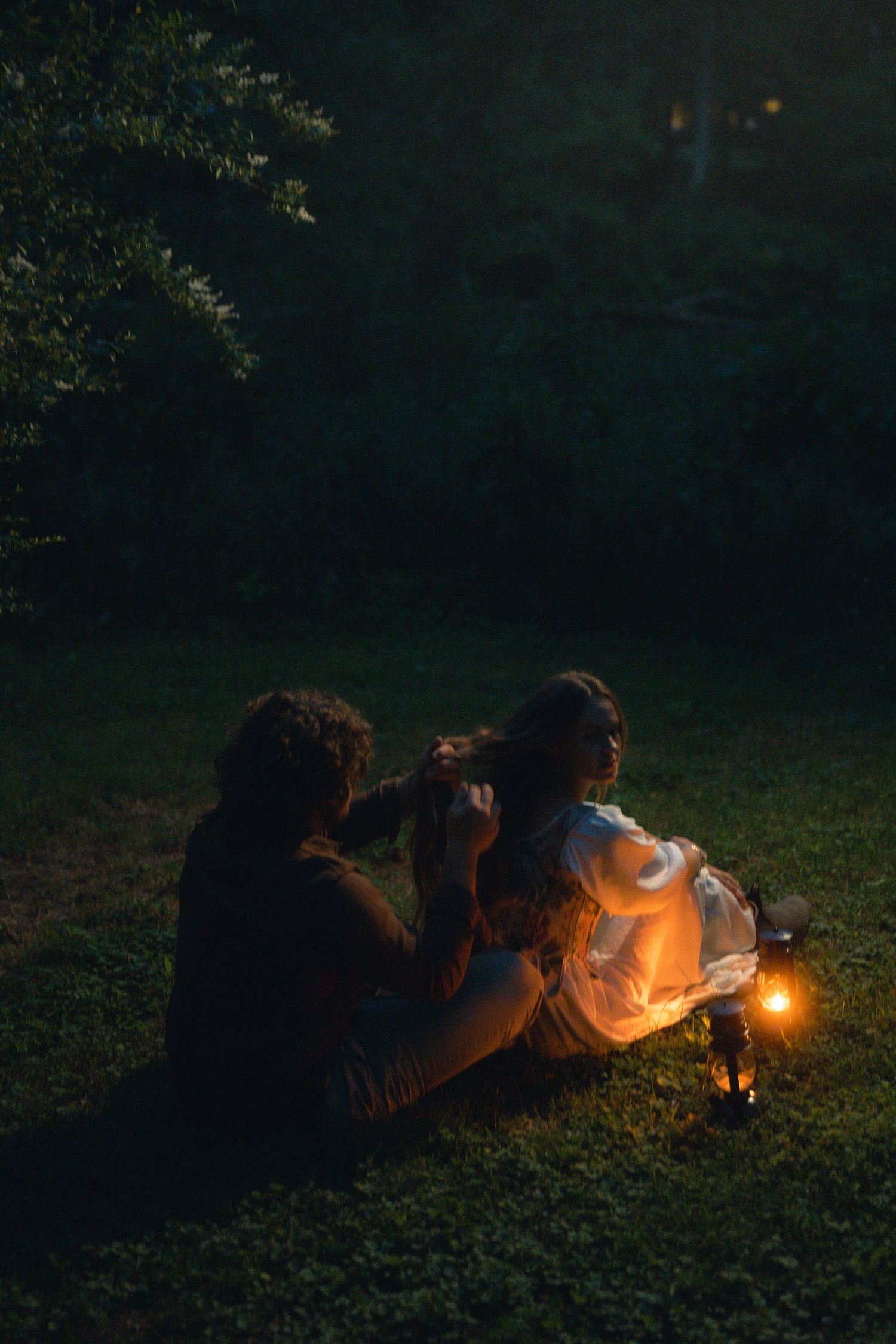 Couple sitting on grass at night illuminated by a warm glowing lantern in a dark garden setting.
