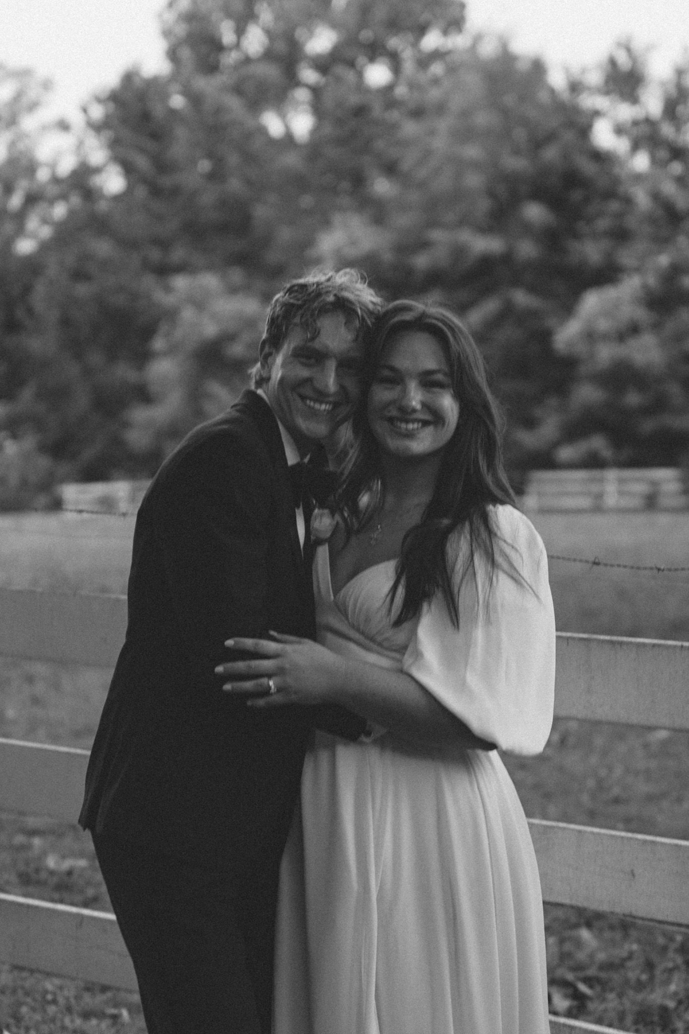 A romantic black and white portrait of a couple embracing by a wooden fence in an outdoor setting.