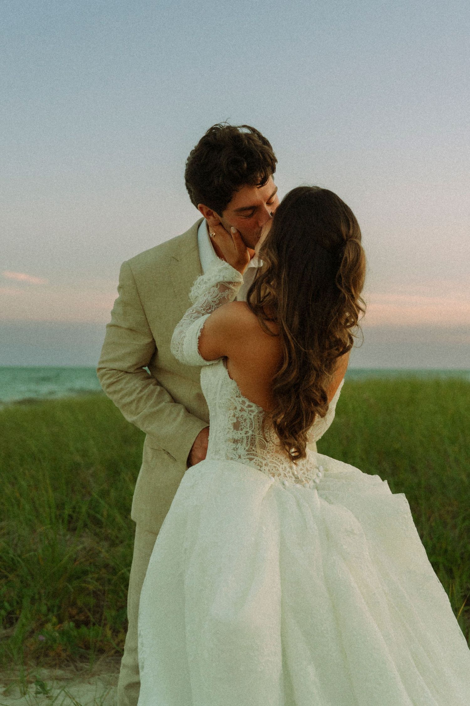 Bride and groom sharing a romantic kiss at sunset on a beach, wearing a white gown and tan suit.