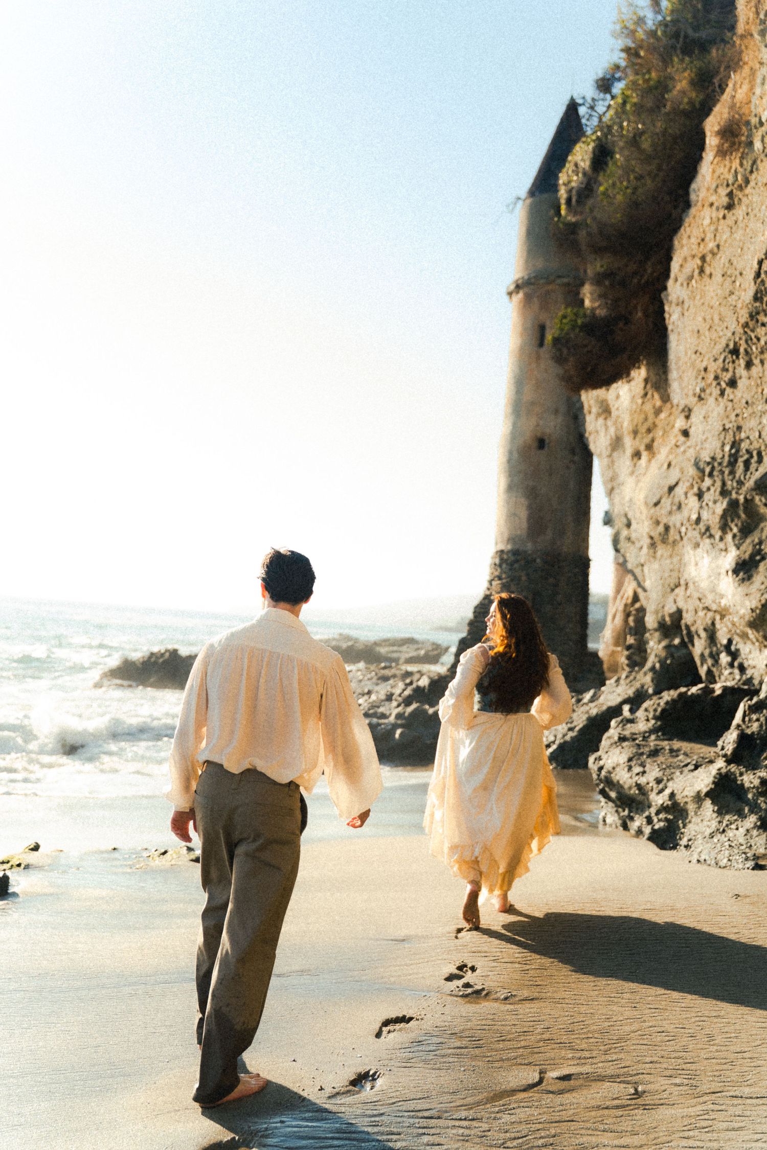 Couple walking along sandy beach near dramatic coastal cliffs.