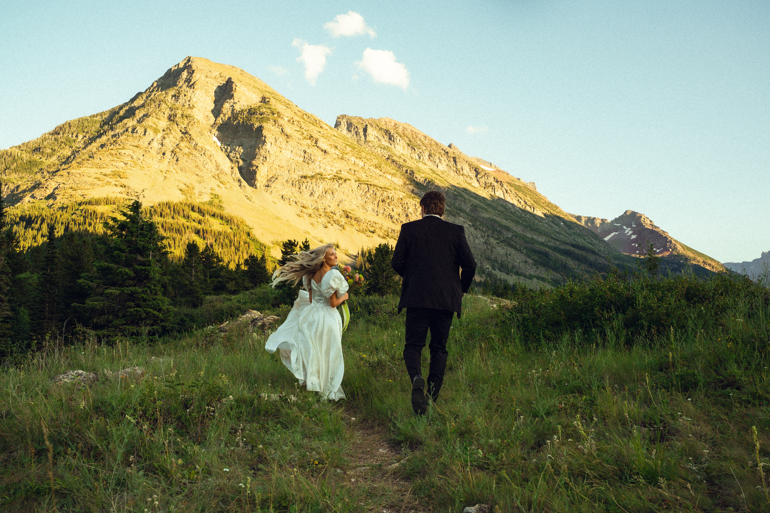 Bride and groom walk hand-in-hand through a mountain meadow trail at golden hour with scenic peaks behind.