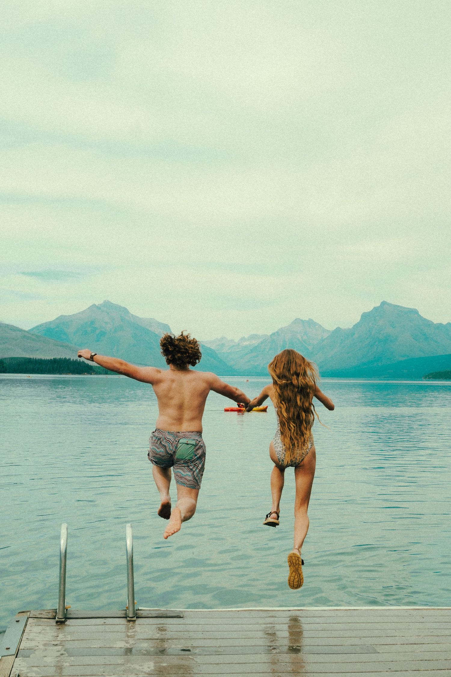 Couple jumping hand-in-hand from a dock into a mountain lake under cloudy skies.