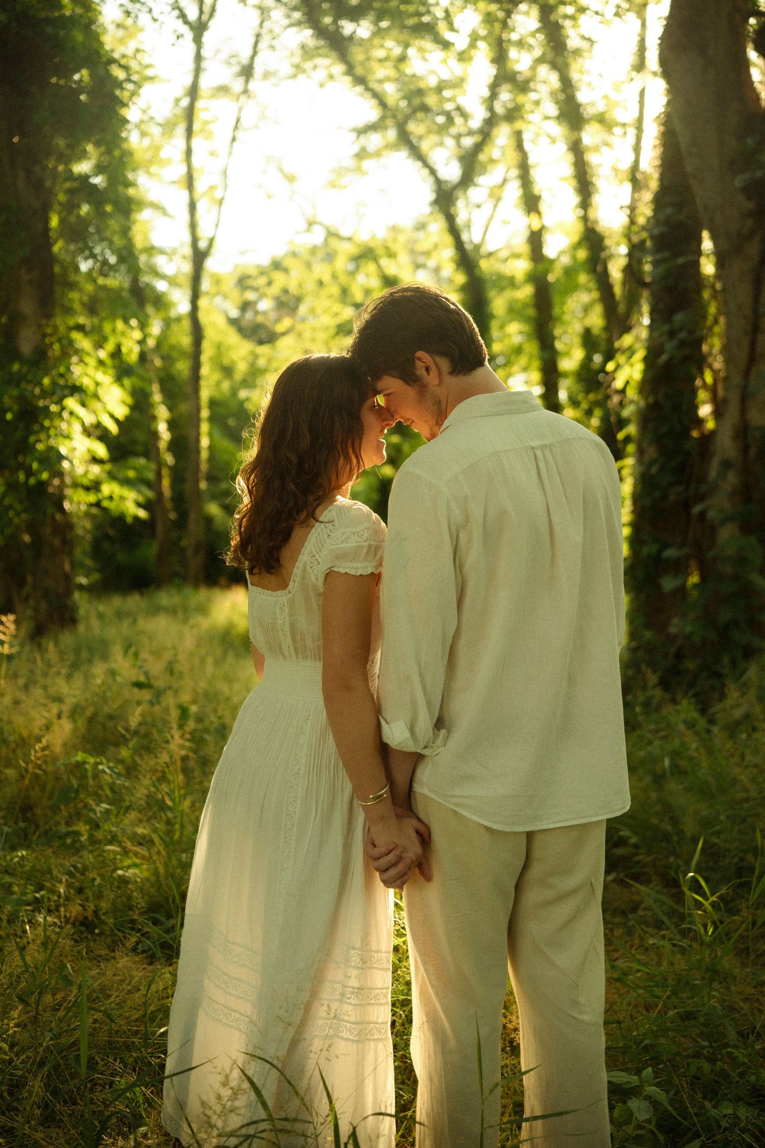 Romantic couple in white outfits holding hands in a sunlit forest, sharing an intimate moment among lush green trees.